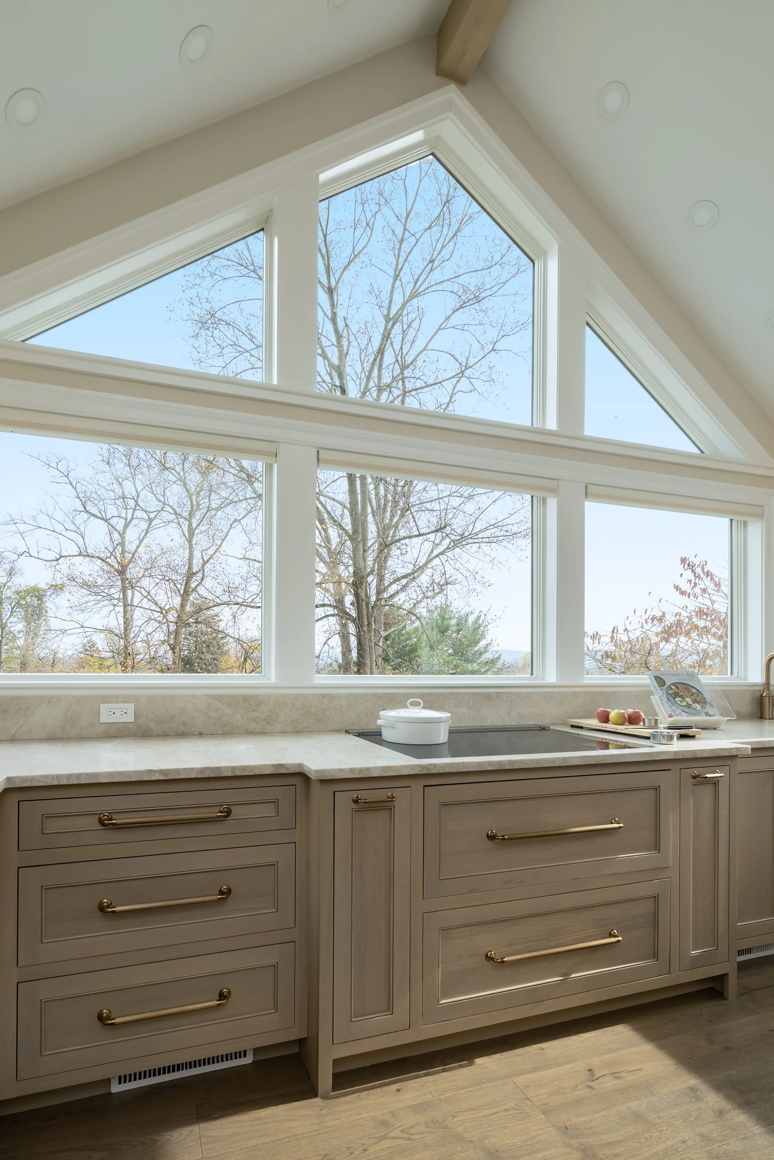 Modern kitchen with beige cabinetry, marble countertop, and large window showing trees outside. Kitchen accessories include a white pot, a cookbook, and some apples.