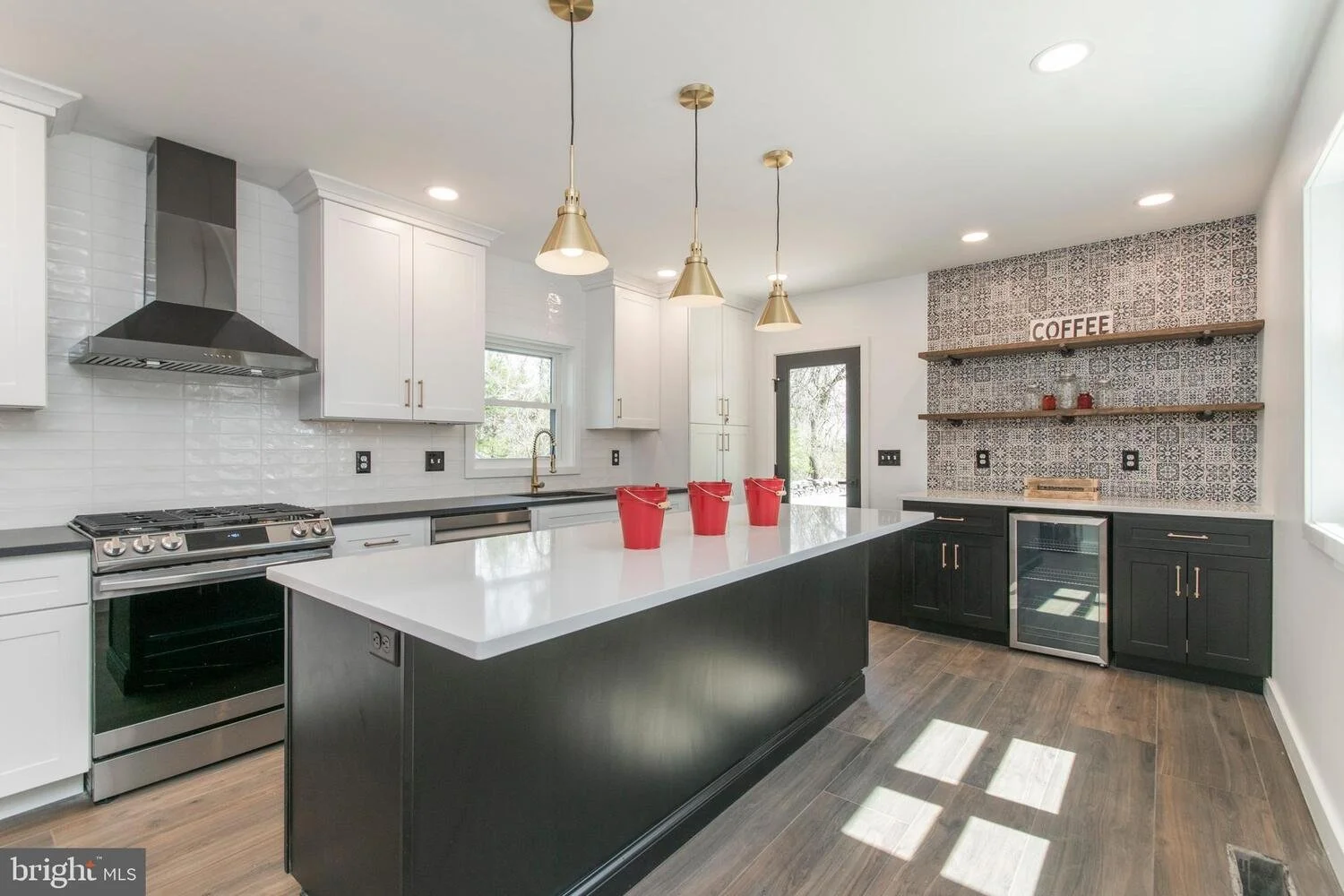 Modern kitchen with white cabinets, black lower cabinets, and a black kitchen island. Features a white countertop, pendant lights, stainless steel oven, and a small wine cooler. Decor includes a coffee sign and decorative shelves on a patterned backsplash.