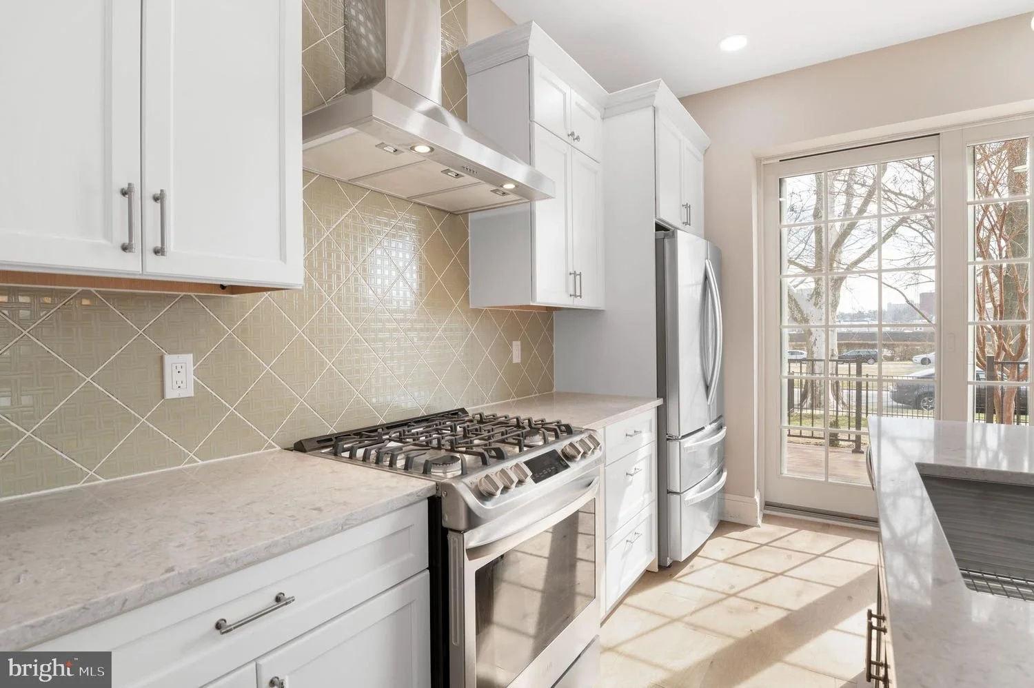 Bright kitchen with white cabinets, granite countertops, beige tiled backsplash, stainless steel stove and dishwasher, refrigerator, and large window door leading outside.