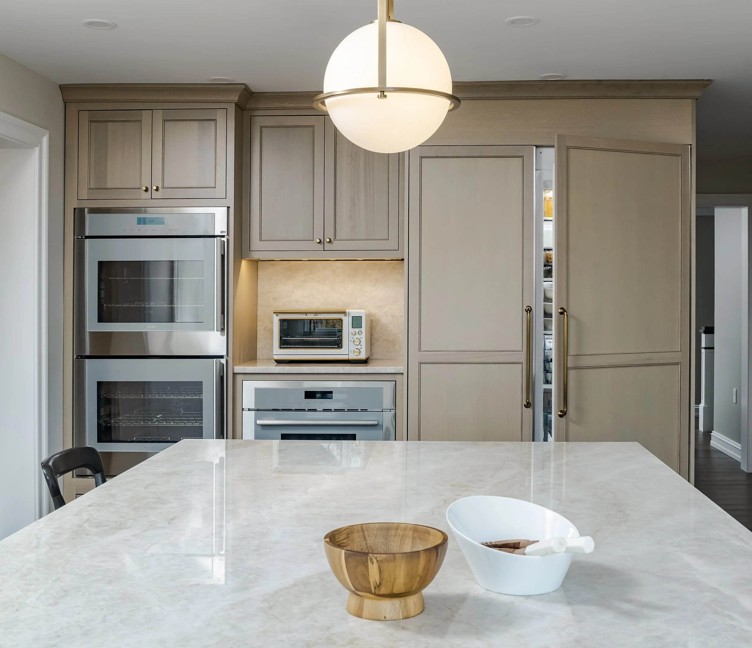 A domestic kitchen with a large white marble island countertop, two bowls (one wooden, one white), and black dining chairs. There are built-in light wood cabinets, a stainless steel oven, a microwave, and a smaller oven on the wall. A modern light fixture hangs from the ceiling.
