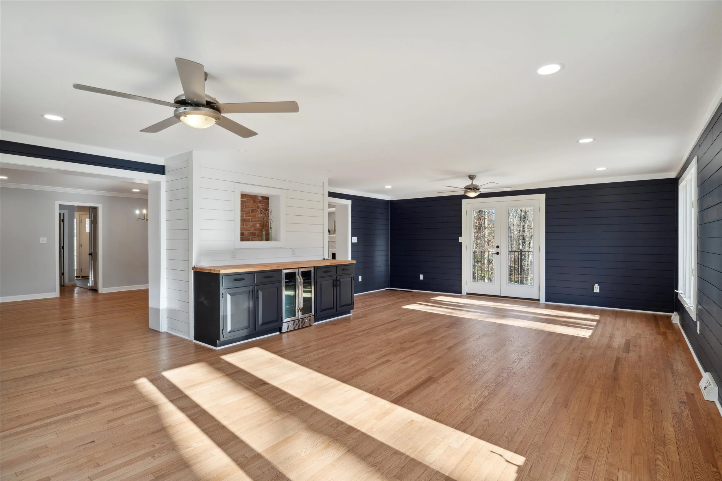 Bright living room with light wood flooring, white ceiling with recessed lights, a ceiling fan, dark blue walls, and large windows with a door leading outside.