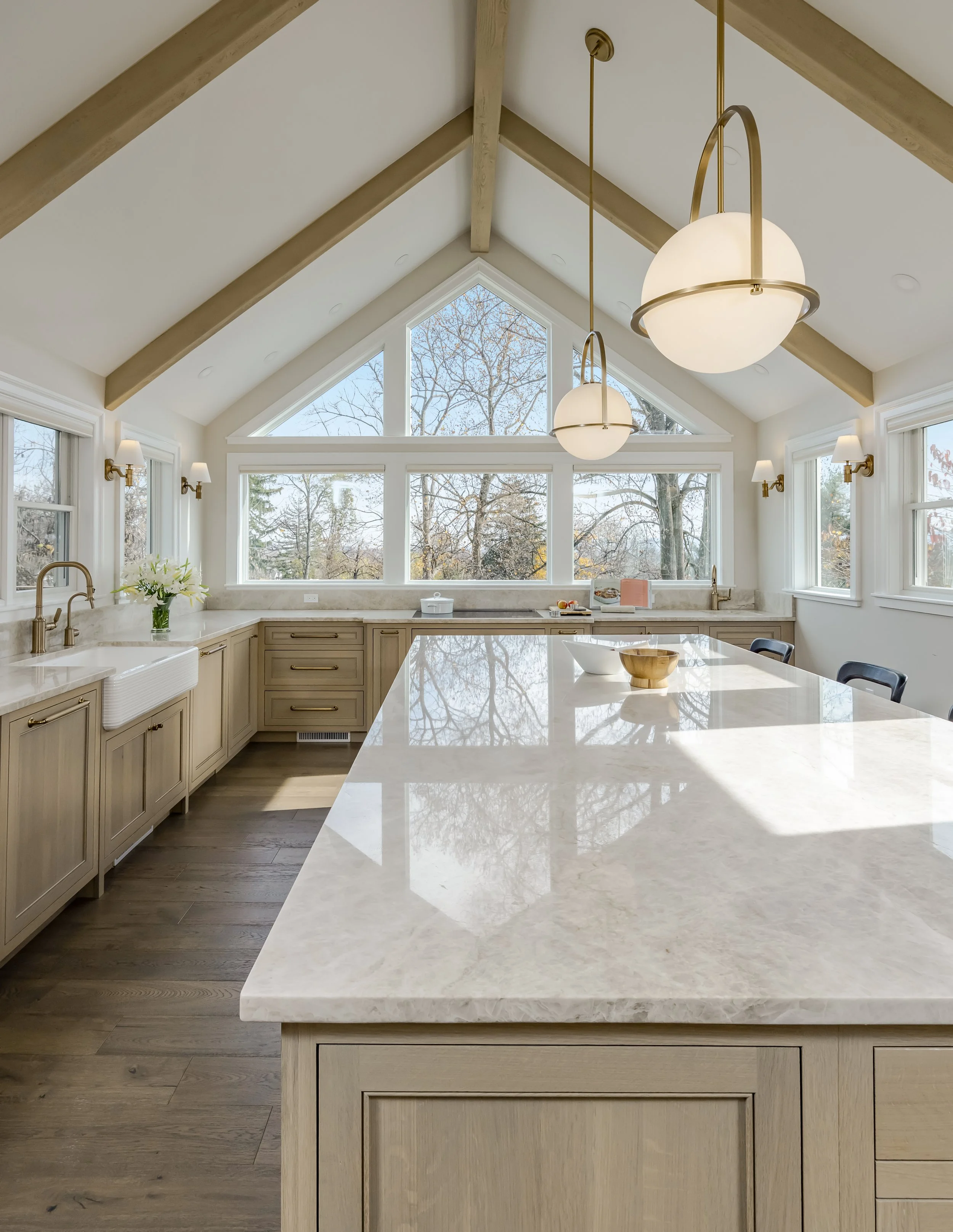 A bright, modern kitchen with large windows and a vaulted ceiling with wood beams. The kitchen features white and beige cabinetry, a marble countertop, and gold and white pendant lights hanging from the ceiling.