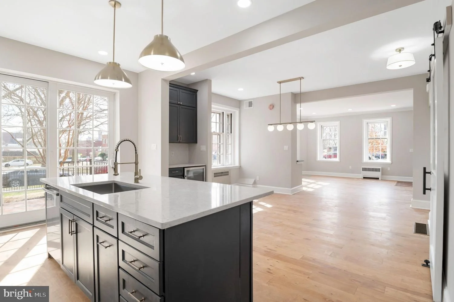 Open kitchen and living room area with hardwood floors, large windows, a kitchen island with a sink, modern pendant light, black cabinets, and bright natural light.