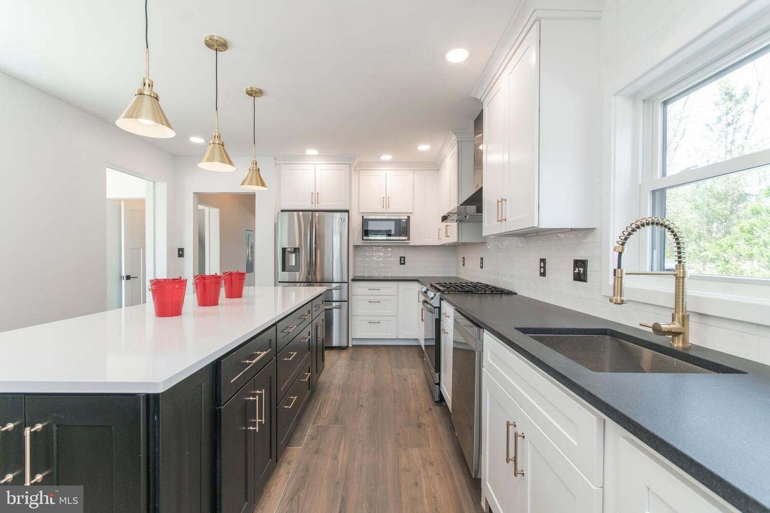 Modern kitchen with white cabinets, contrasting black lower cabinets, stainless steel appliances, gold hardware, black countertops, a large window, and three red buckets on the island.