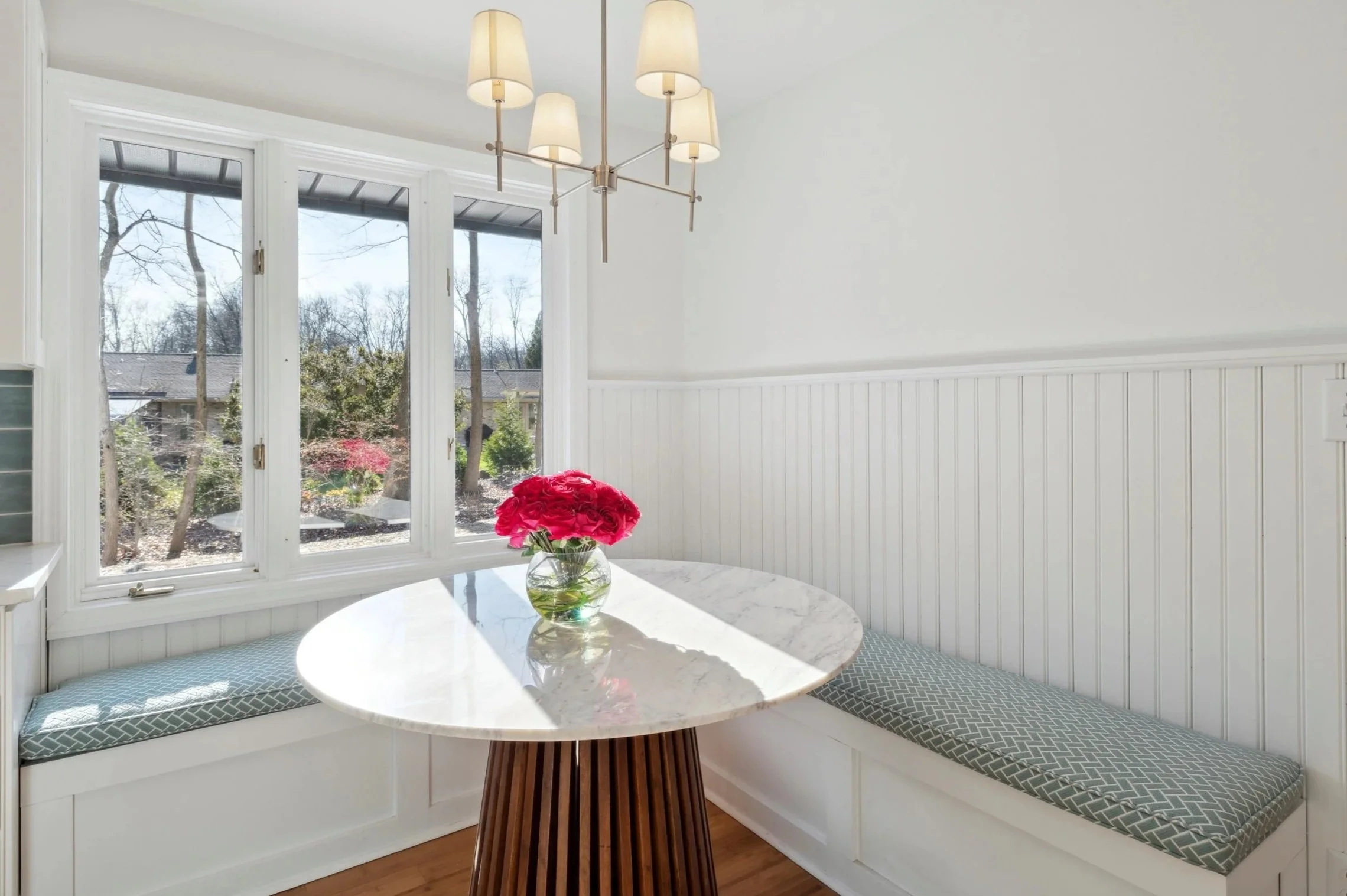 A bright kitchen corner with a curved white subway tile bench, cushioned seating with geometric patterned cushions, a white marble round table, a glass vase with pink flowers, large windows showing trees outside, and a modern chandelier with five shades hanging from the ceiling.