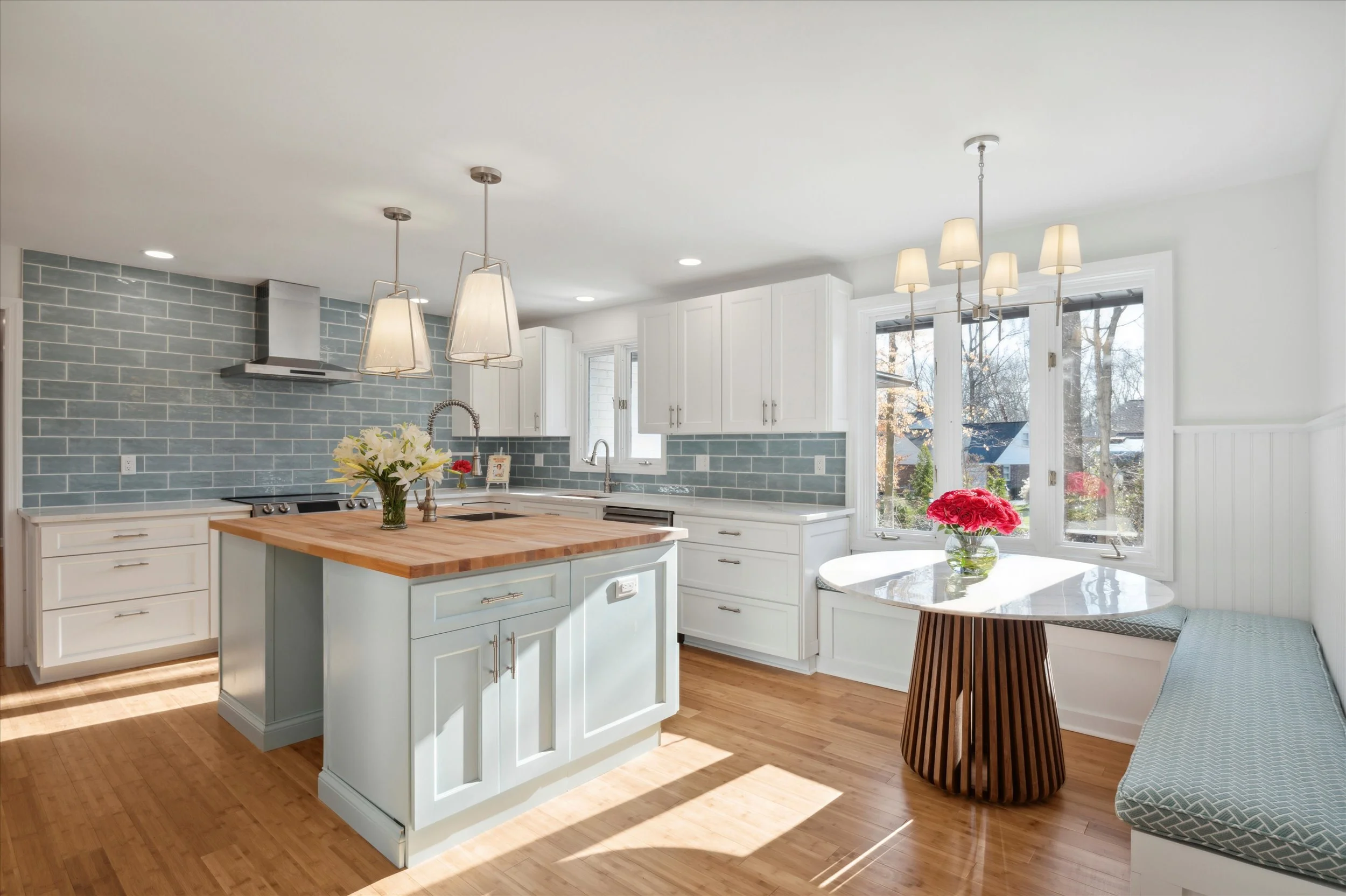 A bright, modern kitchen with white cabinetry, a blue tiled backsplash, and pendant lighting. There is a vase of white lilies and a bowl of pink flowers on the table by the window, with natural light streaming in through large windows.