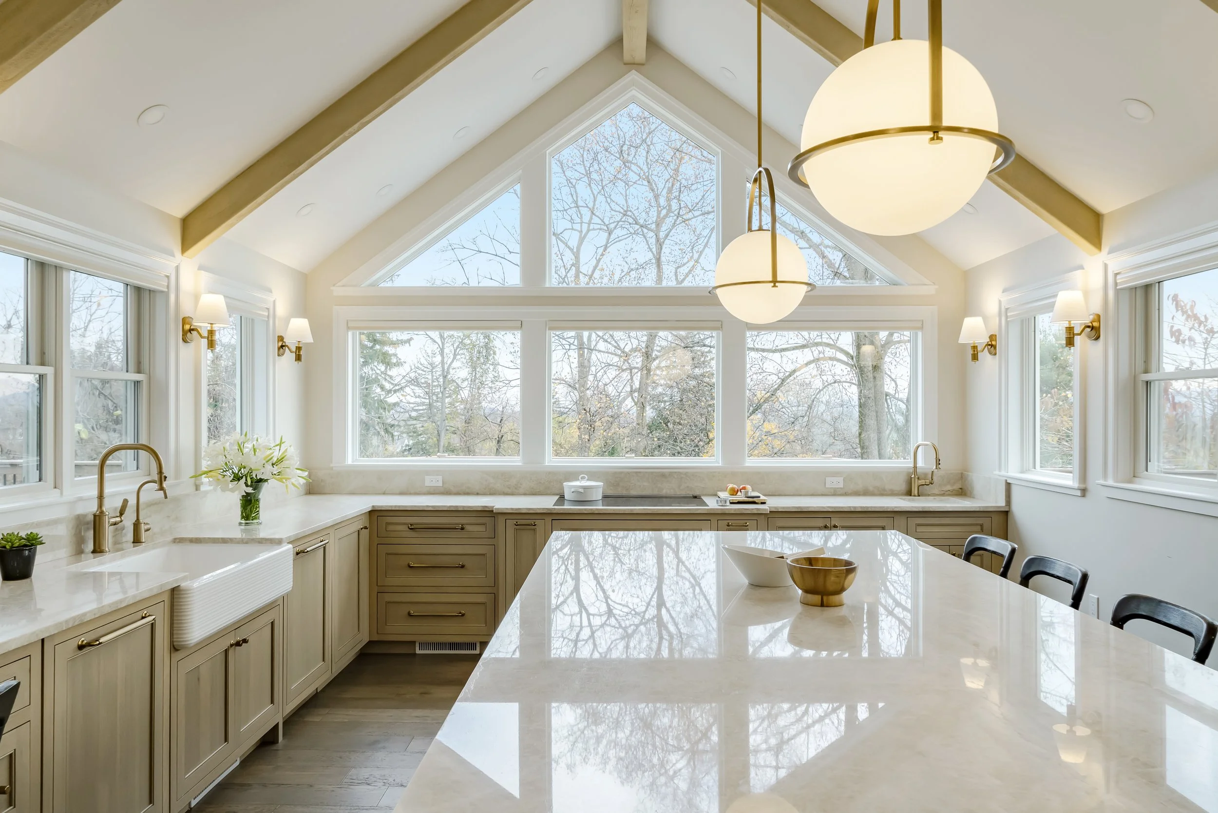 Bright kitchen with large triangular window, beige cabinets, white countertops, gold fixtures, and modern lighting fixtures hanging from the ceiling.