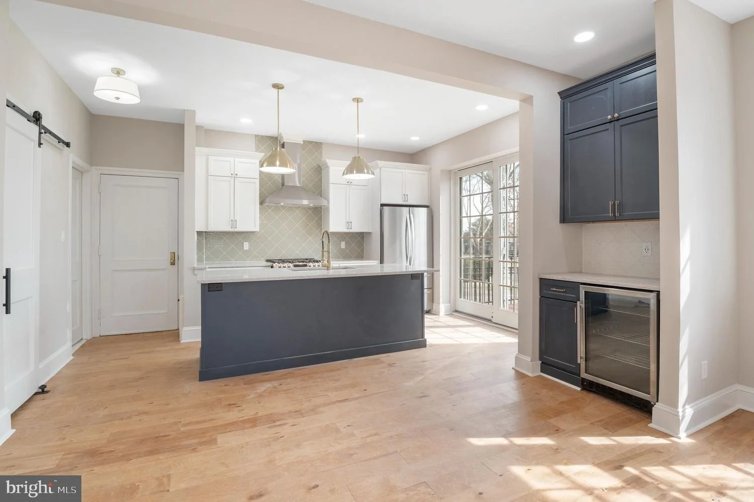 Modern kitchen with white cabinets, dark blue island, stainless steel appliances, light wooden flooring, and large glass doors leading outside.