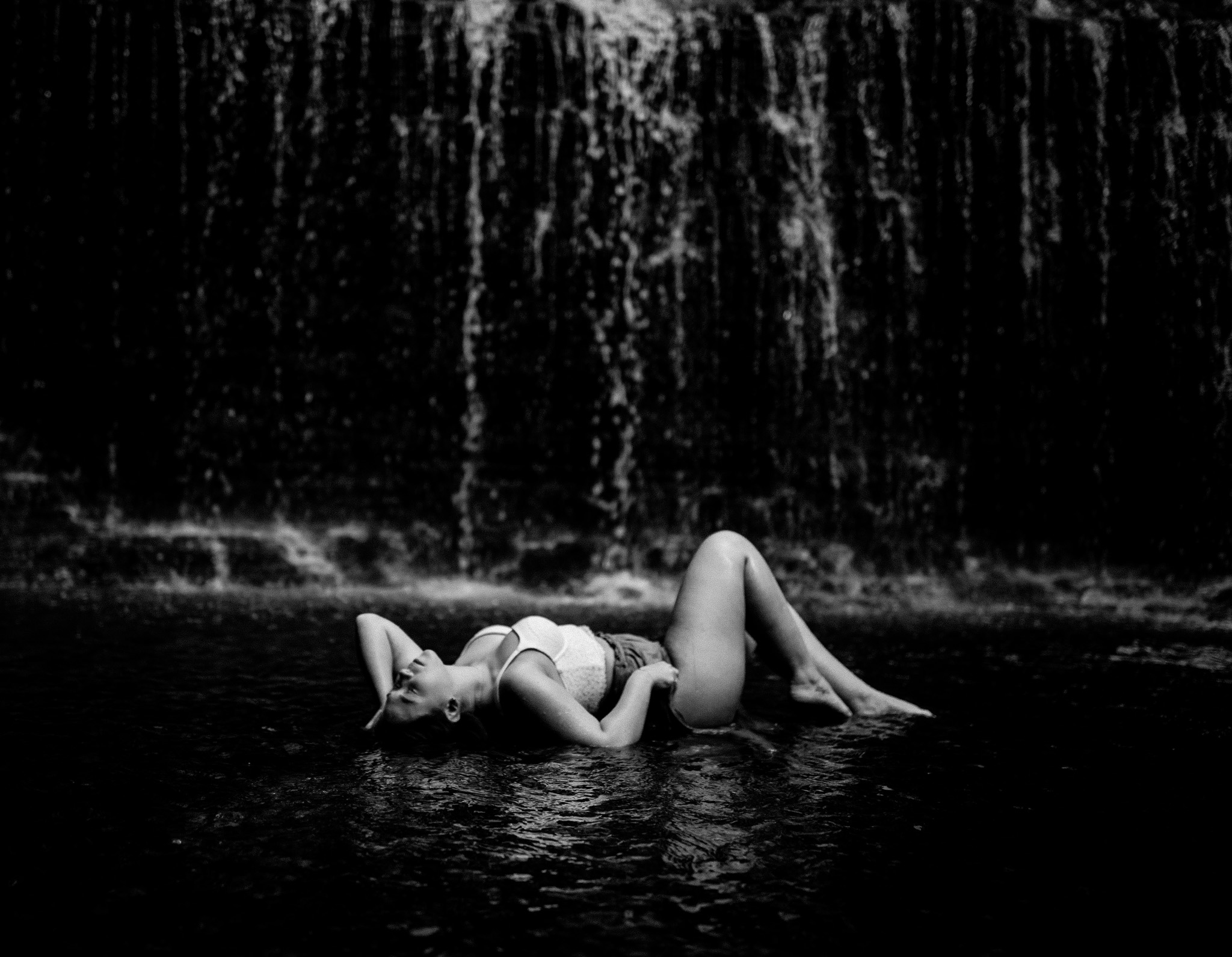 black and white photo of girl laying in water in front of waterfall