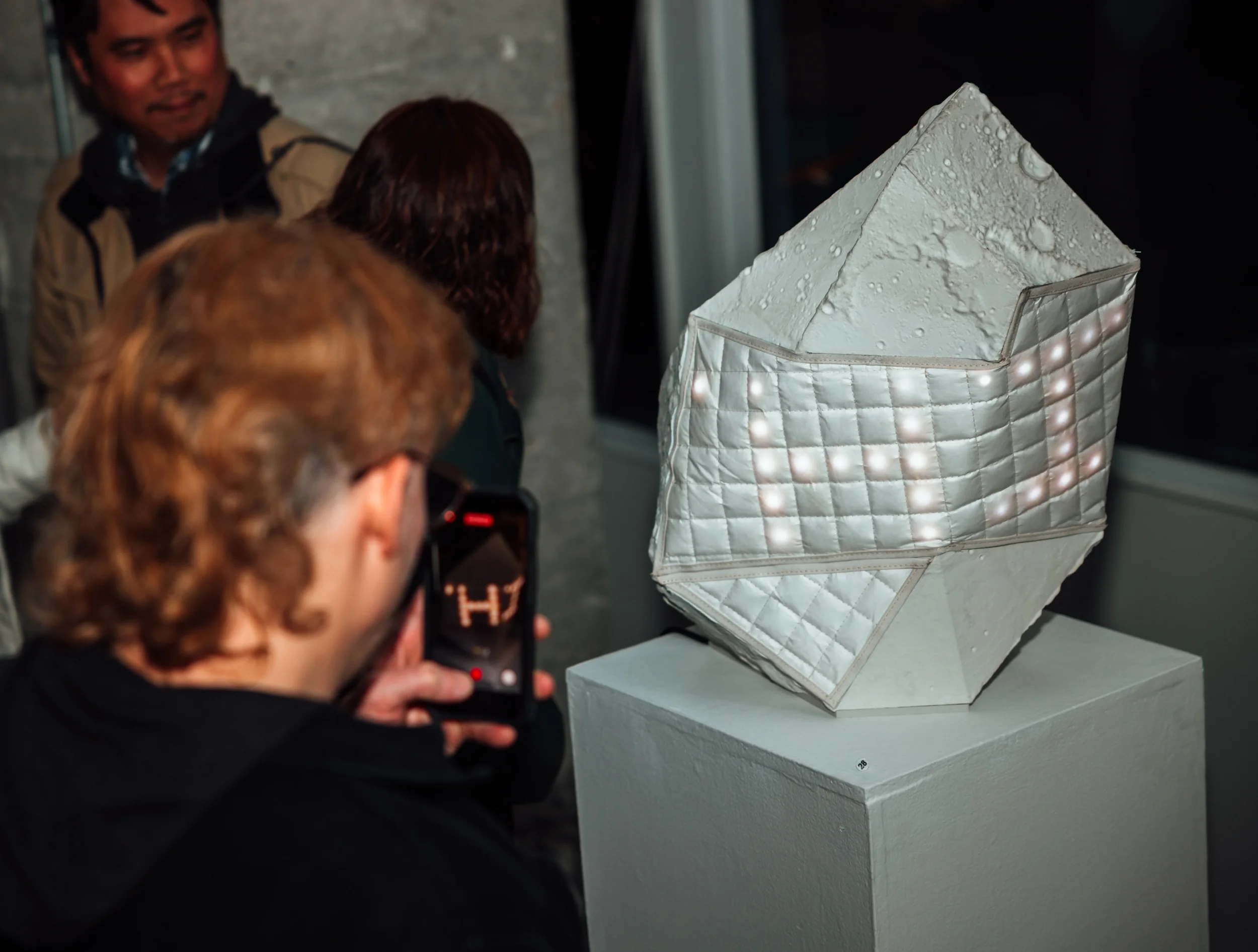 Image of a person photographing the sculpture with their iphone while it's on exhibit in a busy art show.