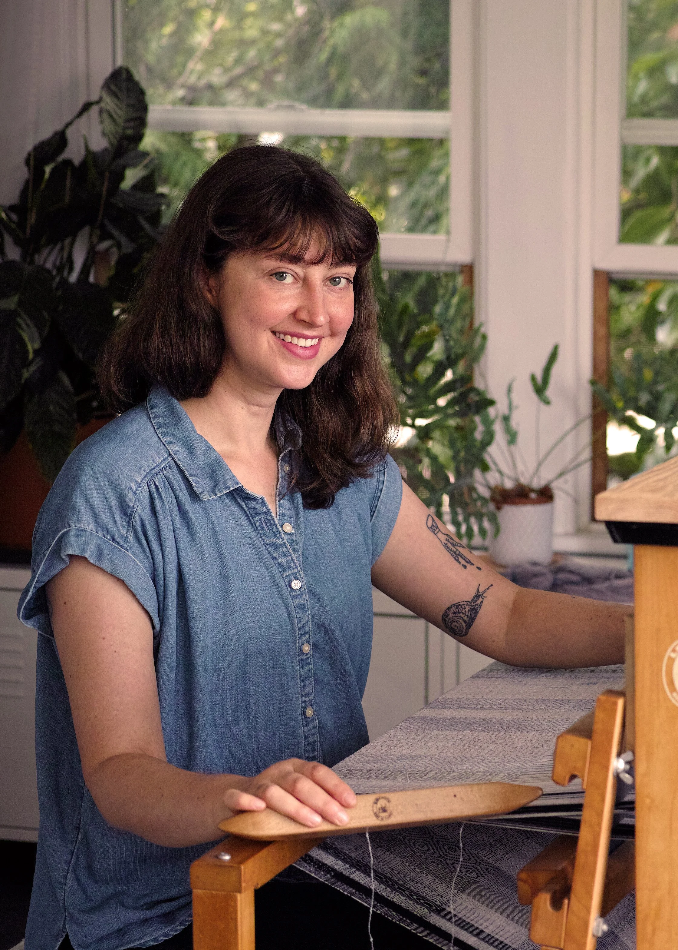 Smiling woman in a denim shirt sits at a weaving loom, with indoor plants and sunlight filtering through windows, conveying a calm, creative atmosphere.
