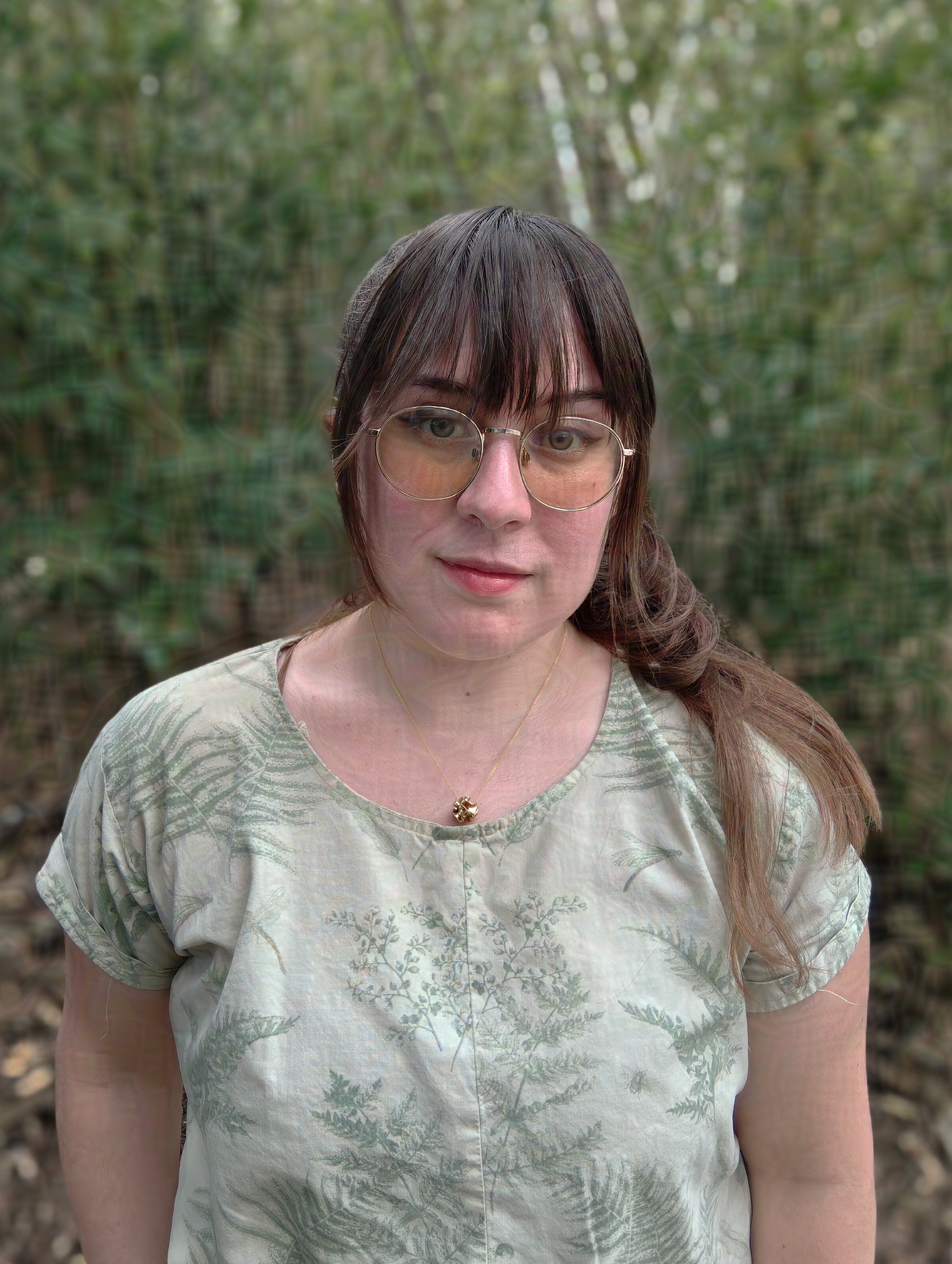 A person with long brown hair and glasses stands outdoors in front of a blurred green leafy background, wearing a leafy-patterned shirt, looking serene.