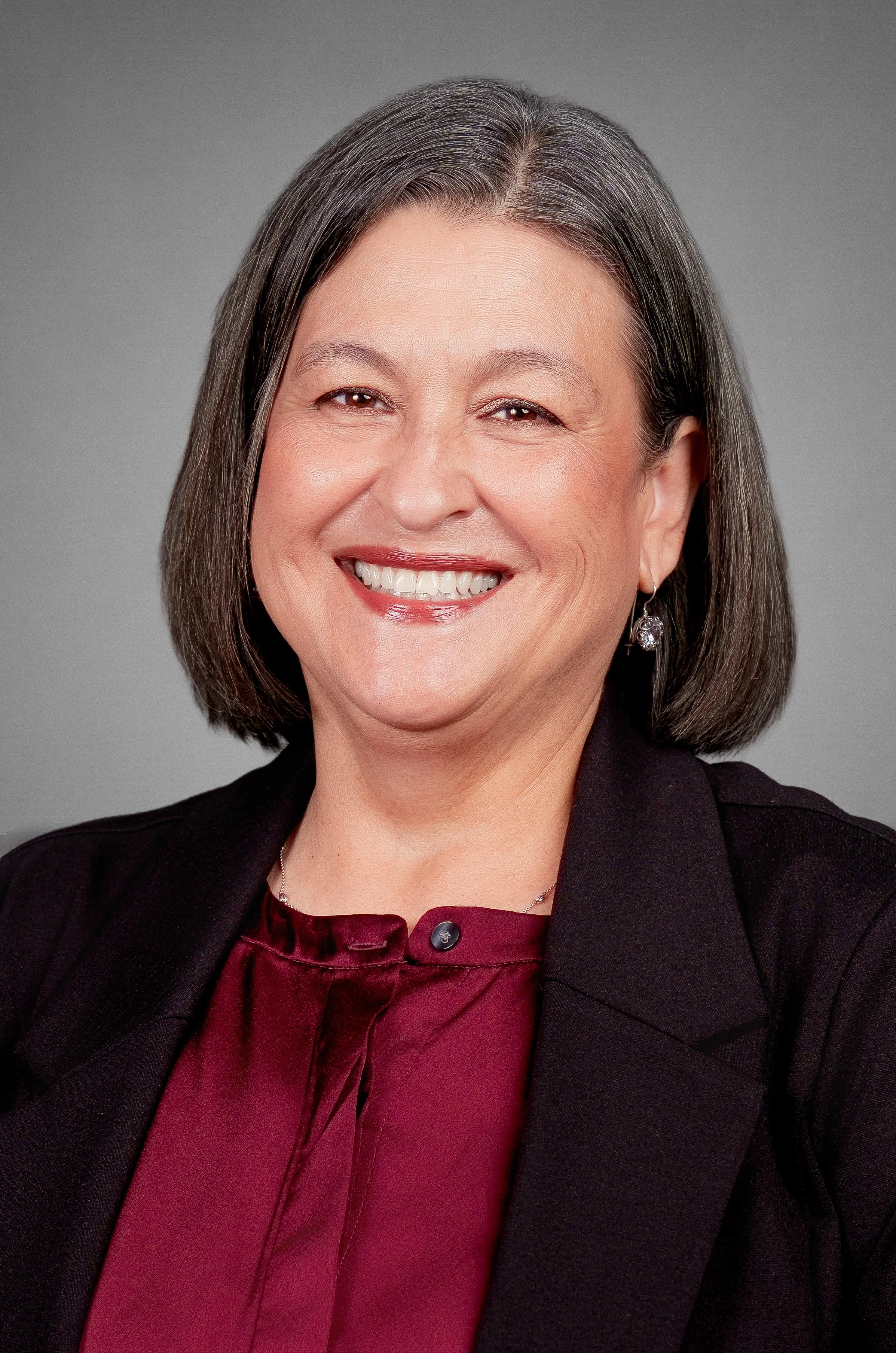 A professional woman with shoulder-length gray hair smiling, wearing a black blazer, maroon blouse with a bow, and earrings, against a gray background.
