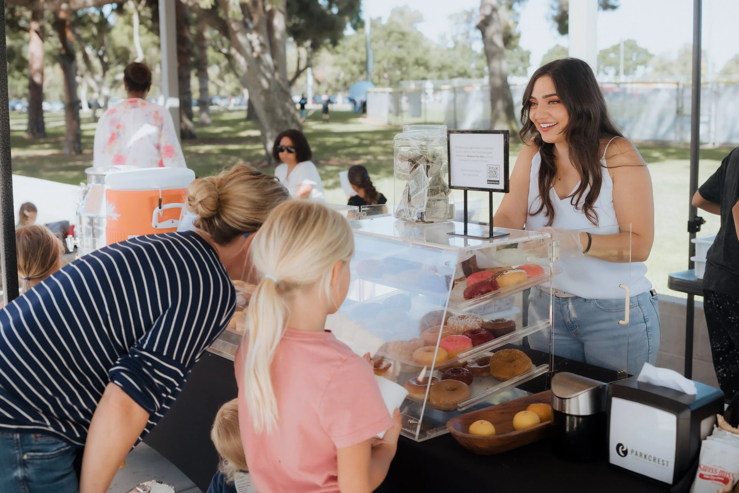 A young woman working at a donut stand, serving customers at an outdoor event on a sunny day, with people and trees in the background.