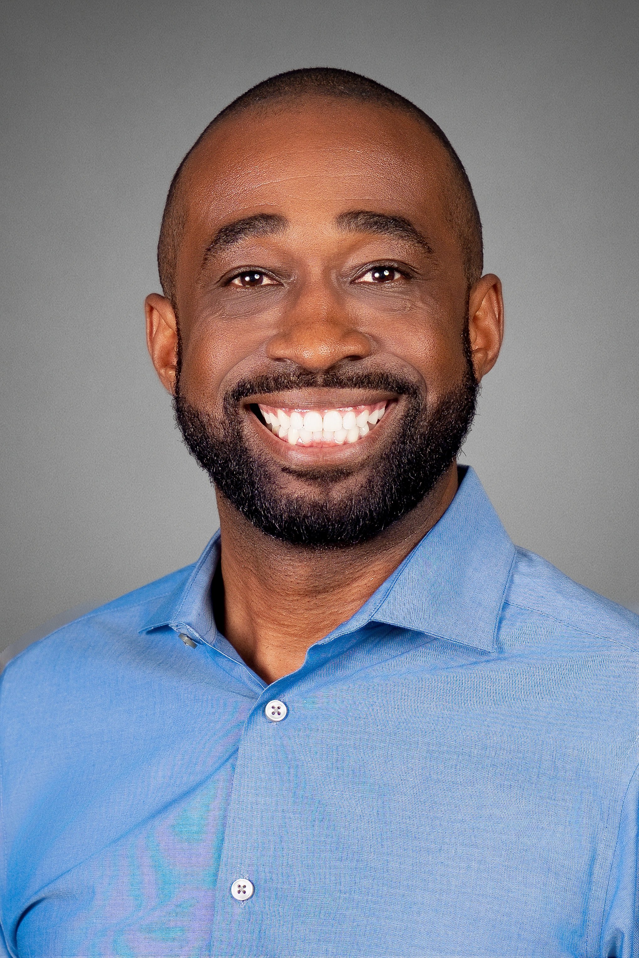 Close-up portrait of an African American man with a broad smile, wearing a light blue collared shirt, with a gray background.
