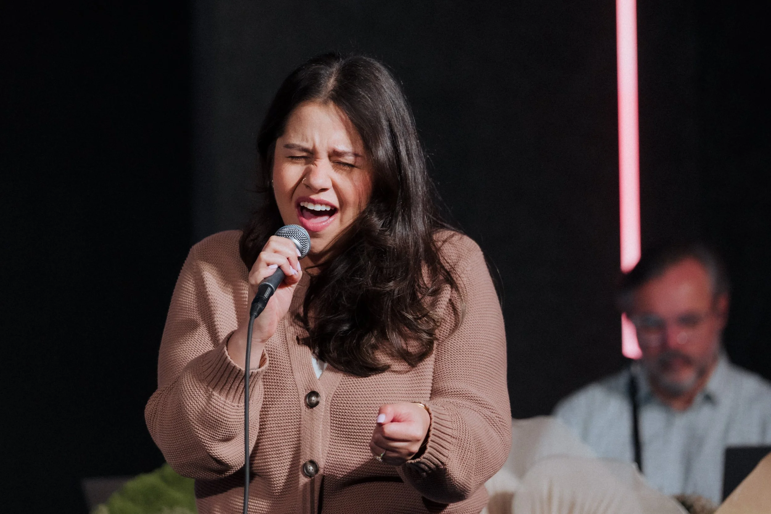 A woman with long dark hair singing passionately into a microphone, wearing a brown cardigan, with other people blurred in the background.