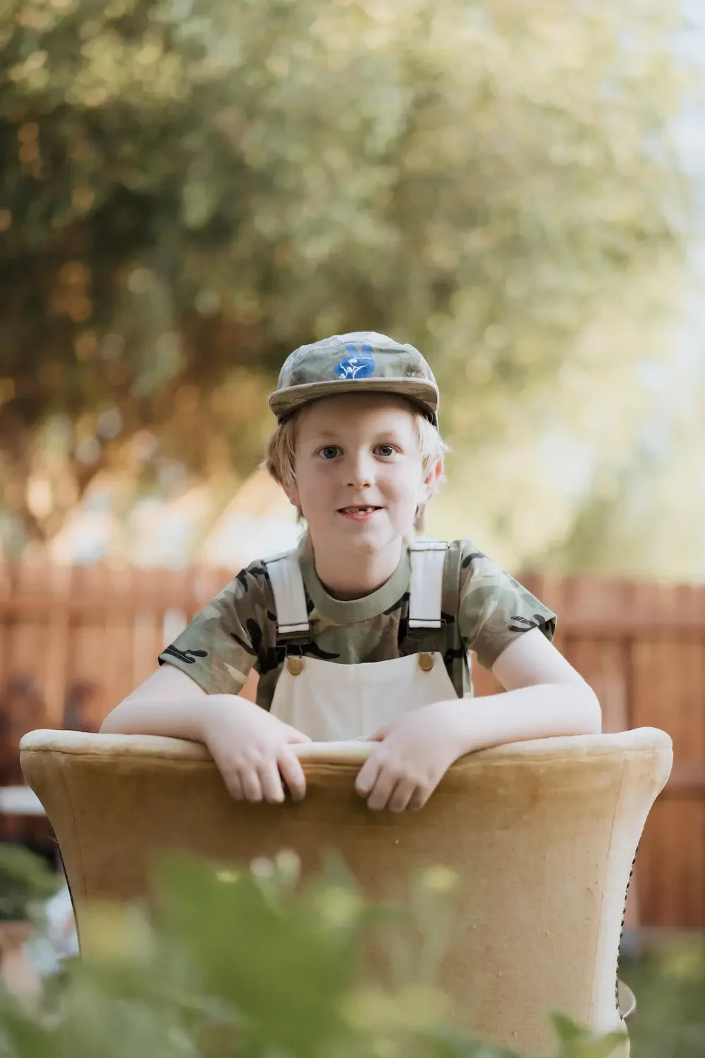 A young boy with blond hair wearing a camouflage t-shirt, white bib, and a child-sized army hat, smiling while leaning on a wooden chair outdoors with a blurred natural background.