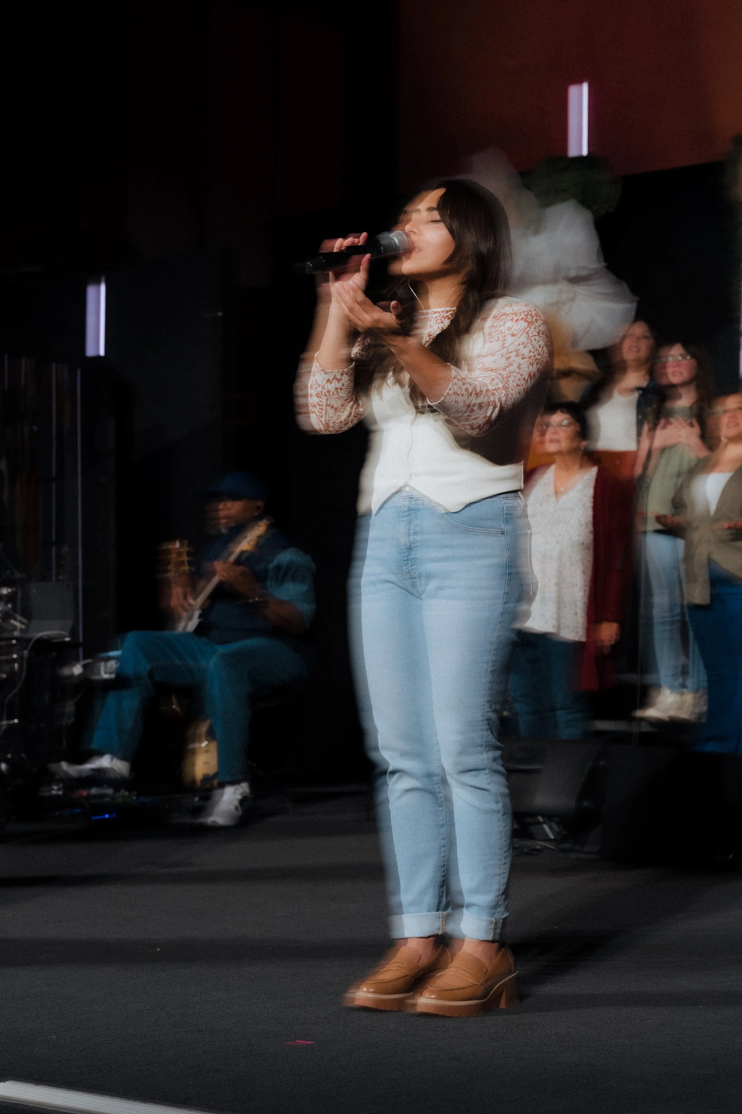 A woman singing into a microphone on stage with a group of people and a musician in the background.