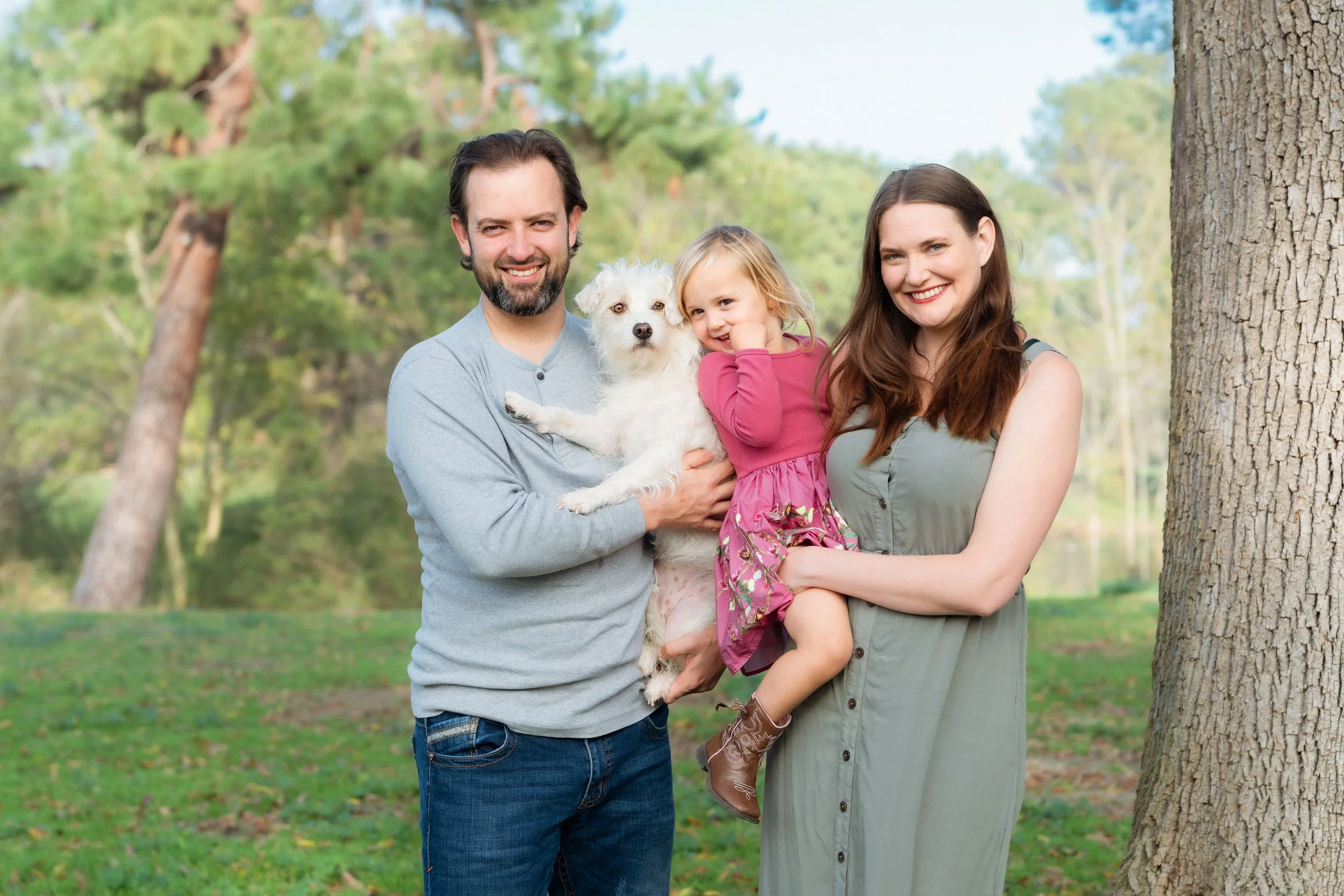 A happy family of four, including a man, woman, young girl, and a dog, standing outdoors in a park with trees and greenery in the background.