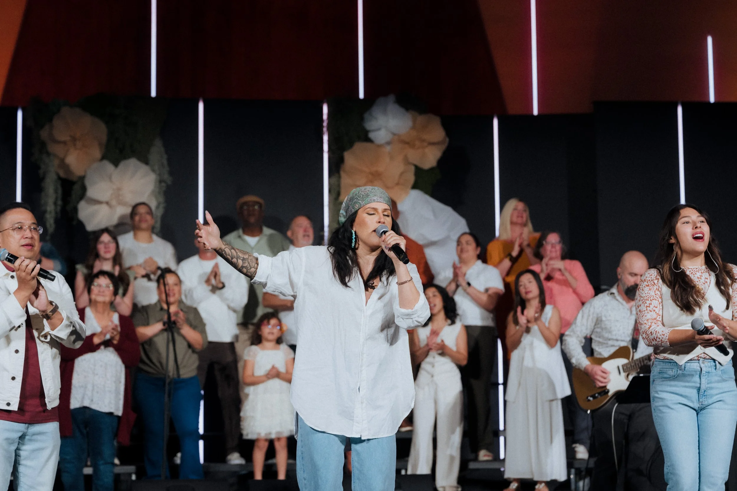 A group of people singing on a stage, with some holding microphones and one person playing the guitar, in a concert or worship setting.