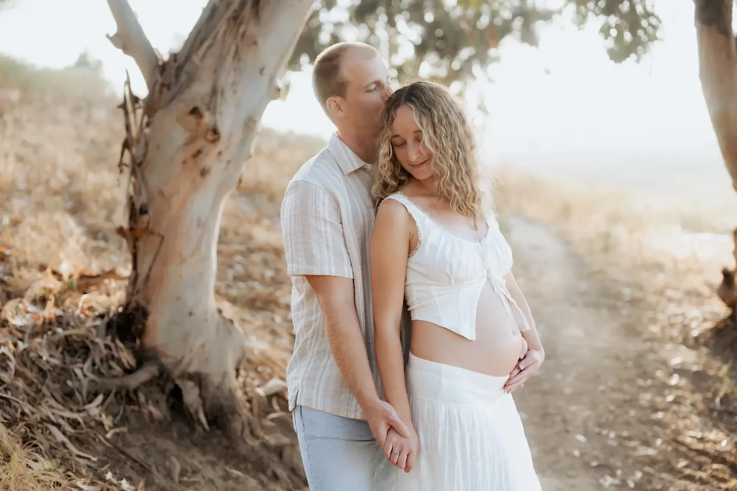 A pregnant woman in a white dress being kissed on the forehead by a man in a light-colored shirt, standing under a tree on a dirt path at Gum Grove in Seal Beach.