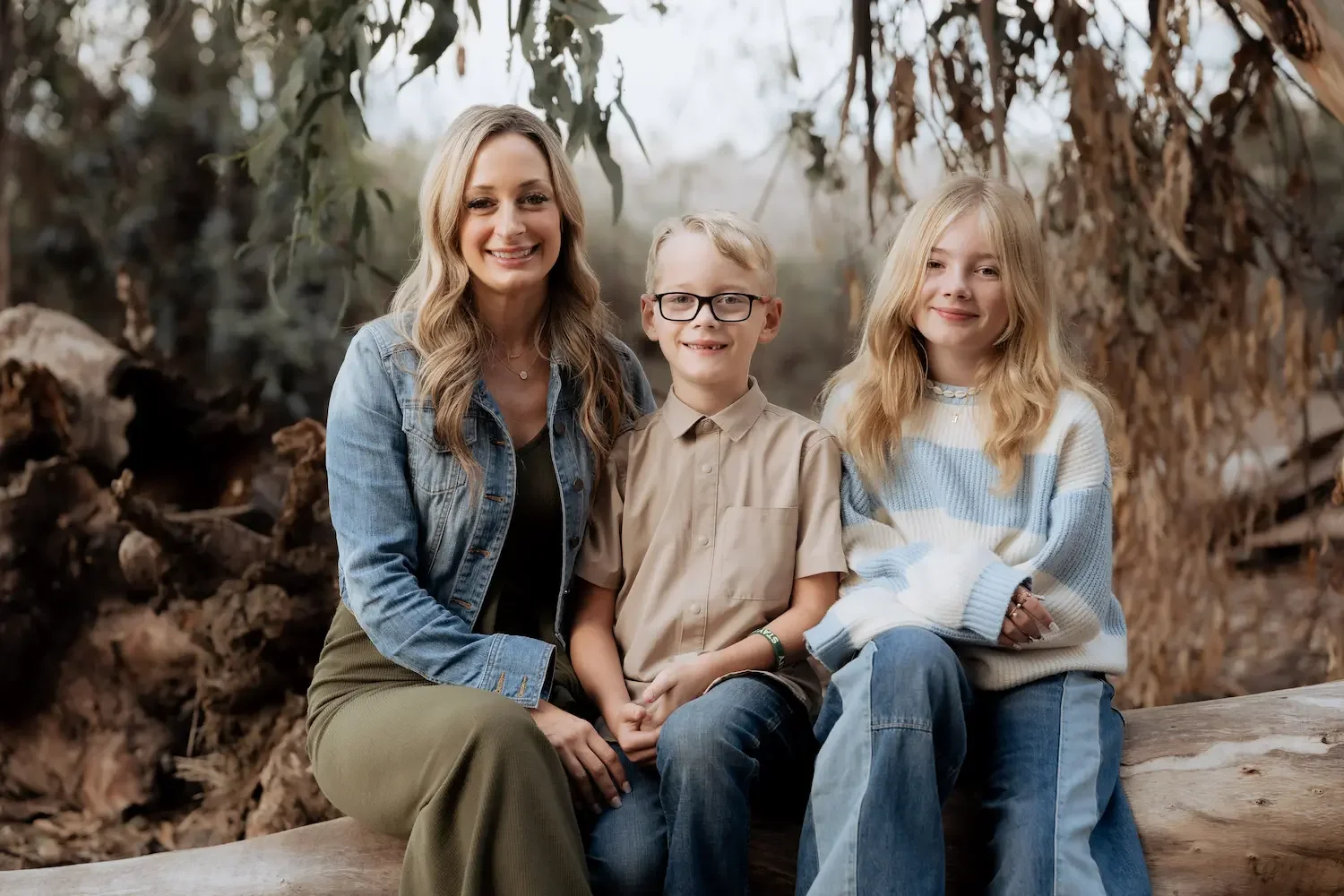 A mother and two children sitting on a log outdoors in Gum Grove Seal Beach, smiling at the camera.