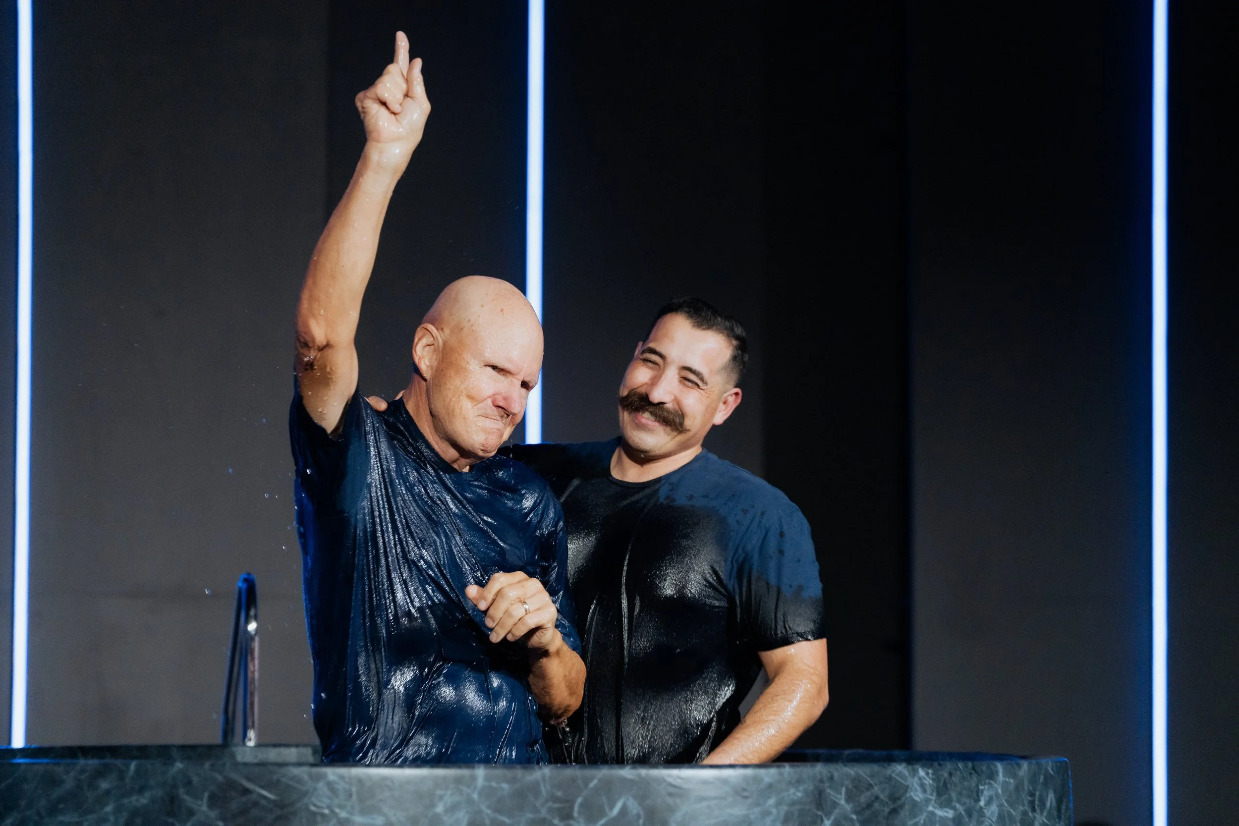 Two men baptizing each other in a baptismal pool, one with a serious expression and the other smiling, both wet and wearing dark shirts, in a modern setting with dark walls and blue vertical lights.
