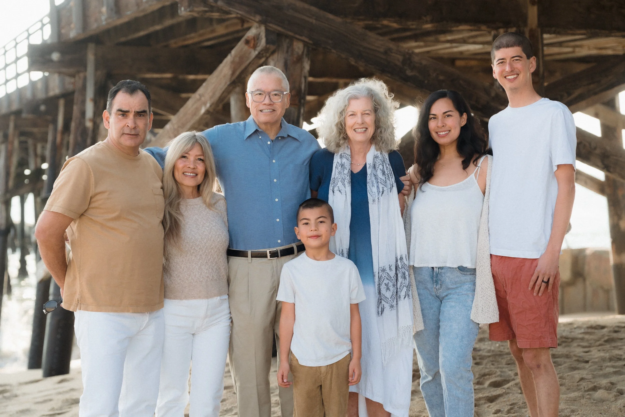 A multi-generational family standing together under a wooden pier on a beach, smiling at the camera.