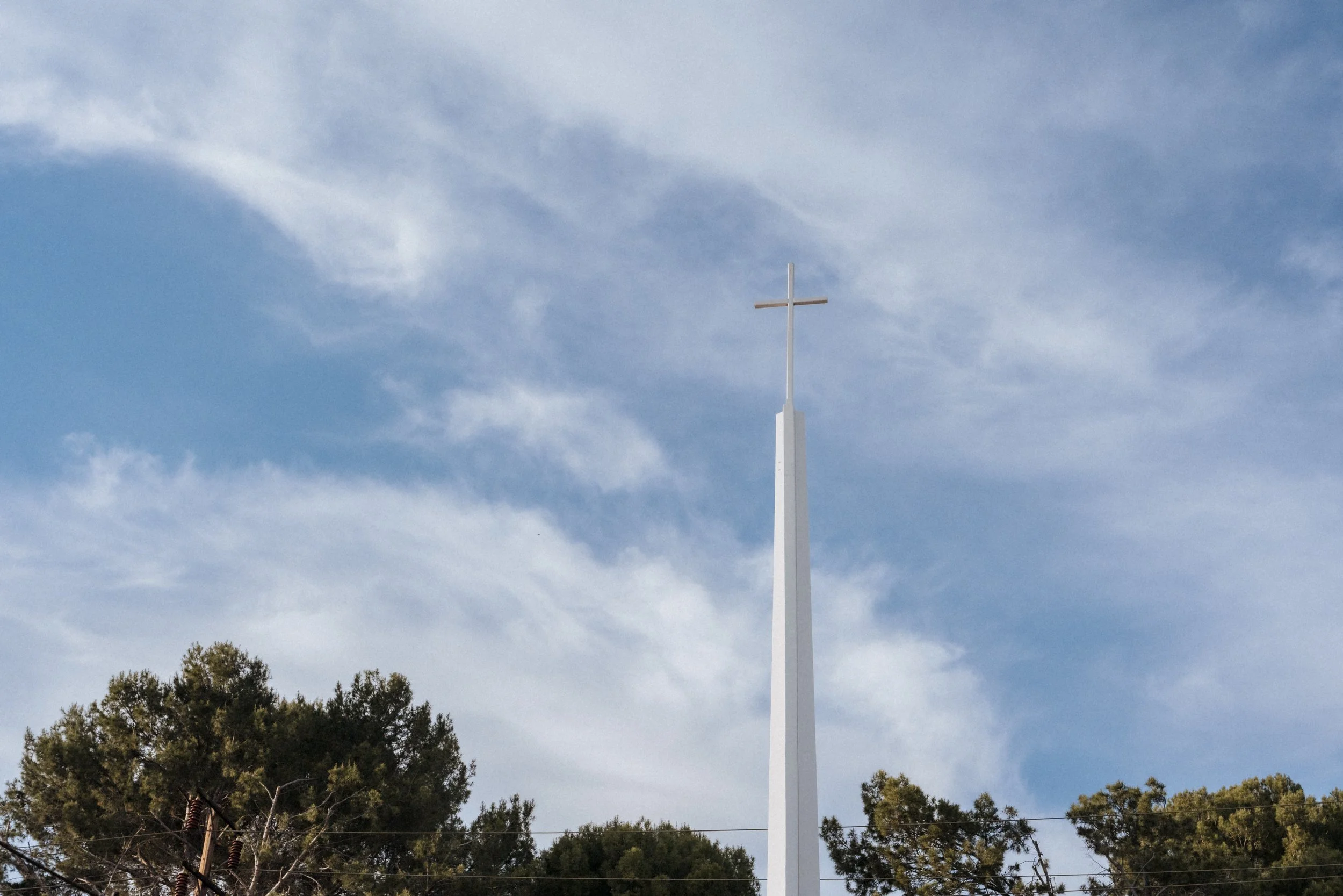 A tall white church steeple with a cross on top against a partly cloudy sky, with trees at the bottom of the image.