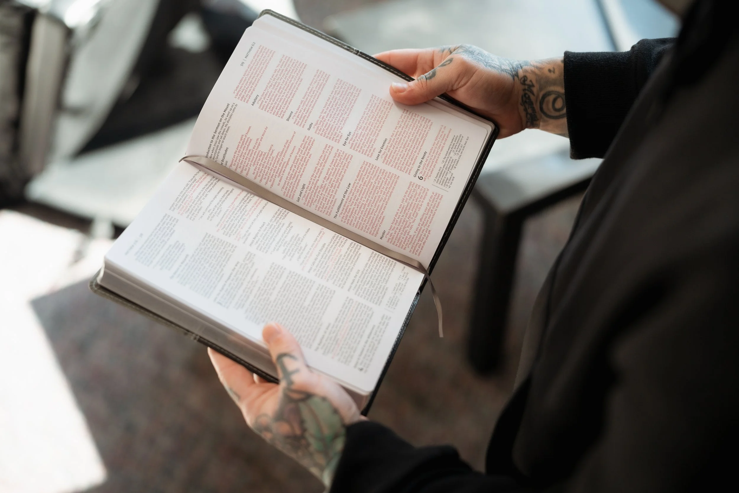 A person with tattoos on their hands holding an open book or Bible, reading it in a well-lit indoor setting.