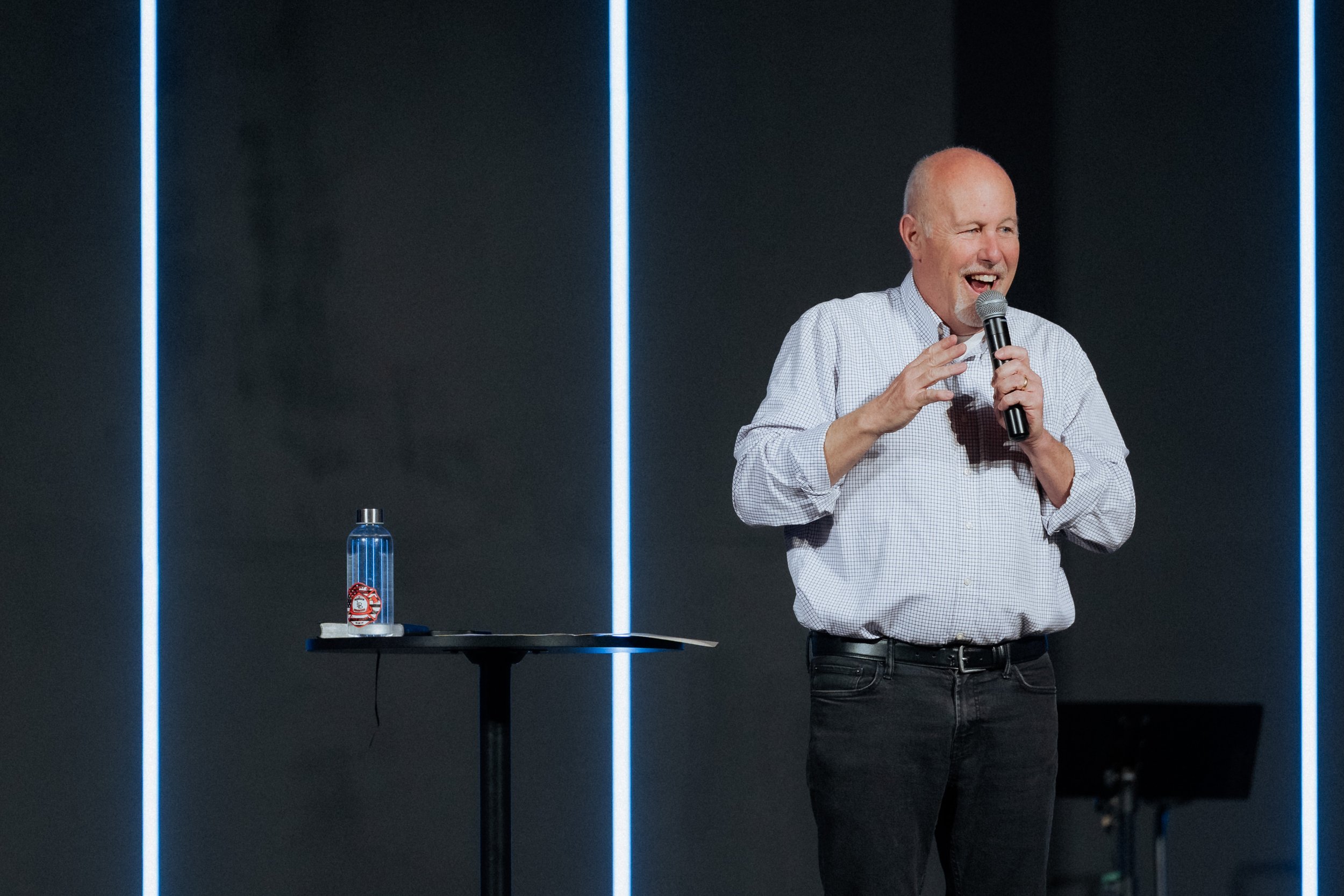 A bald man in a checkered shirt holding a microphone and speaking on stage, with a water bottle on a high table next to him. The background is dark with vertical illuminated lines.