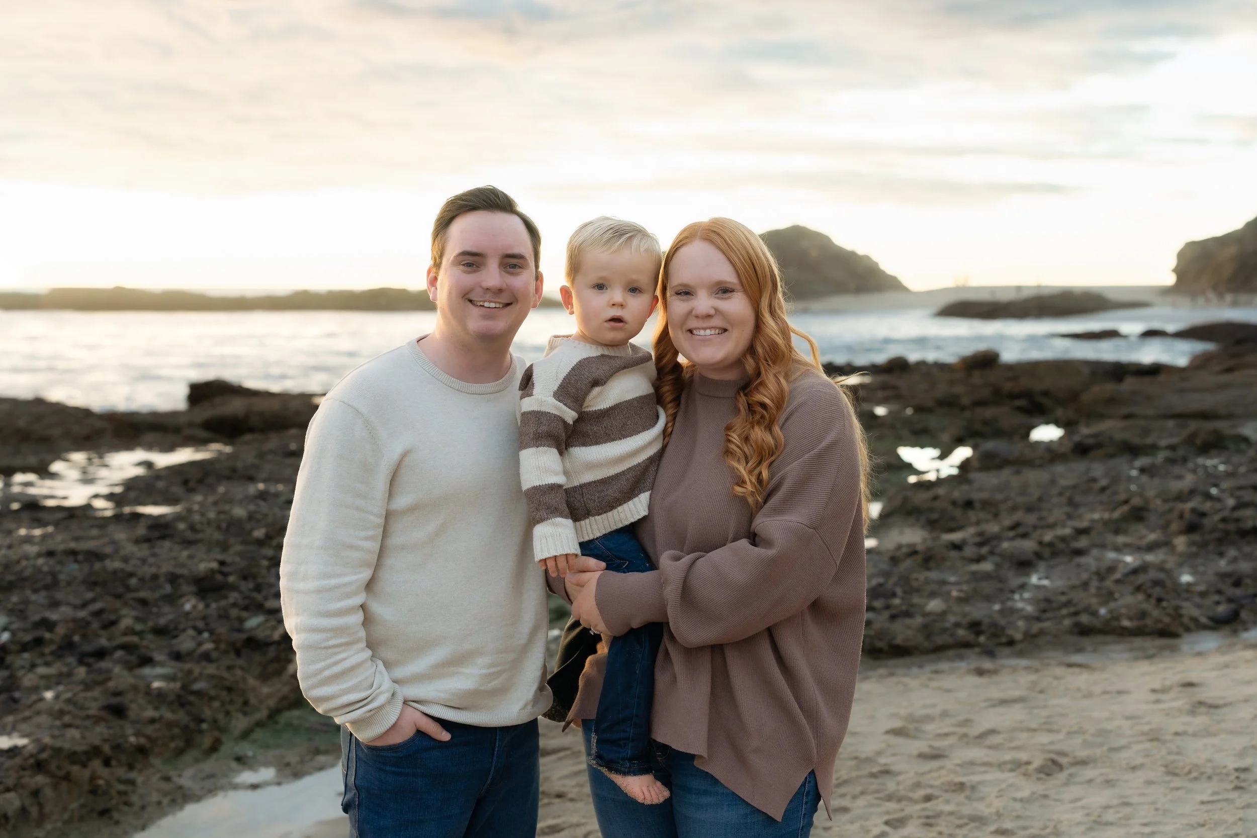 A family of three on a rocky beach during sunset, smiling at the camera. The father has dark hair and is wearing a light-colored sweater. The mother has long, curly red hair and is wearing a brown oversized sweater. The young boy, between them, has b