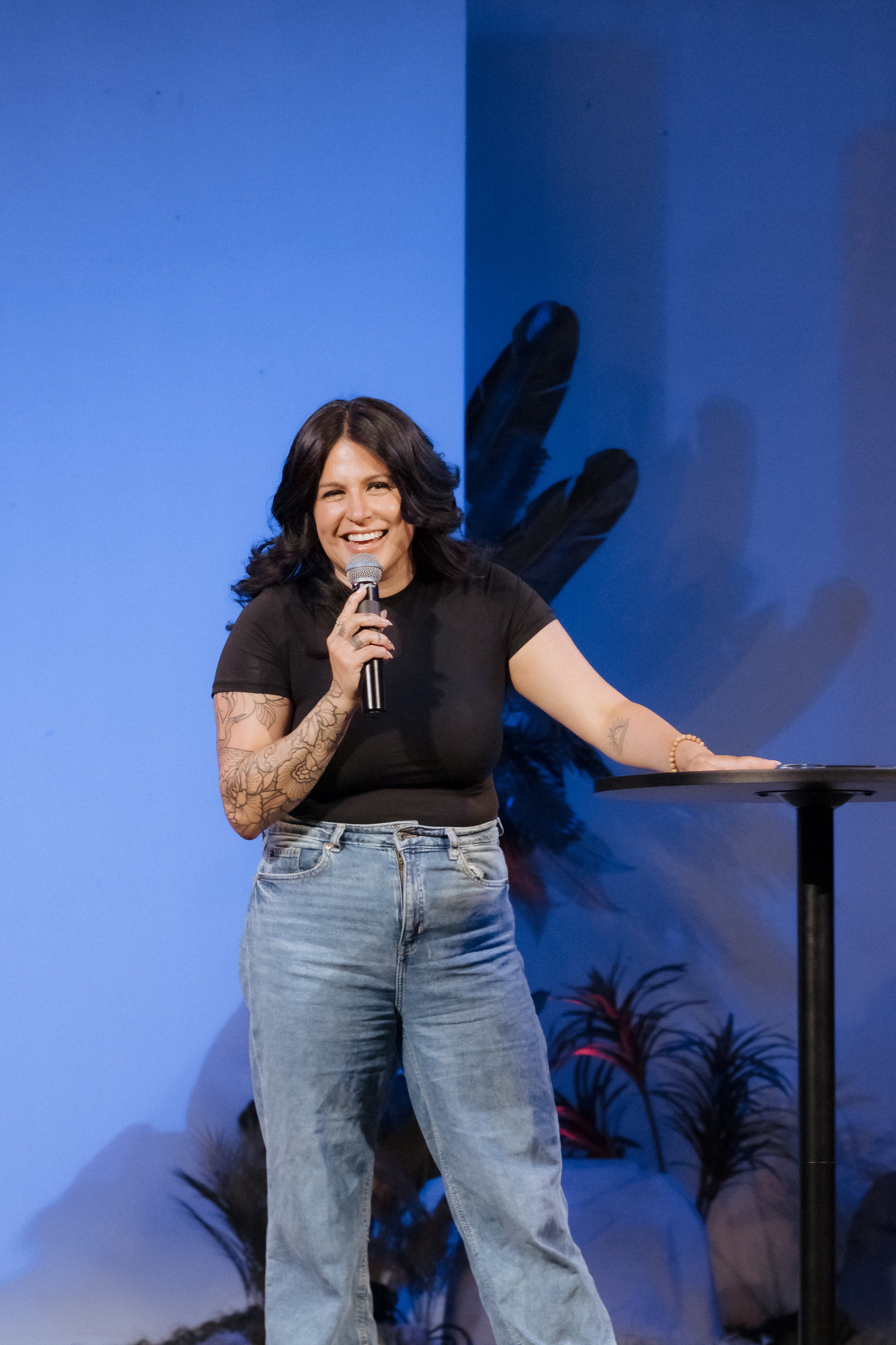 A woman with dark hair, tattoos on her arm, wearing a black t-shirt and jeans, holding a microphone and smiling on stage with a blue background and plant decorations.