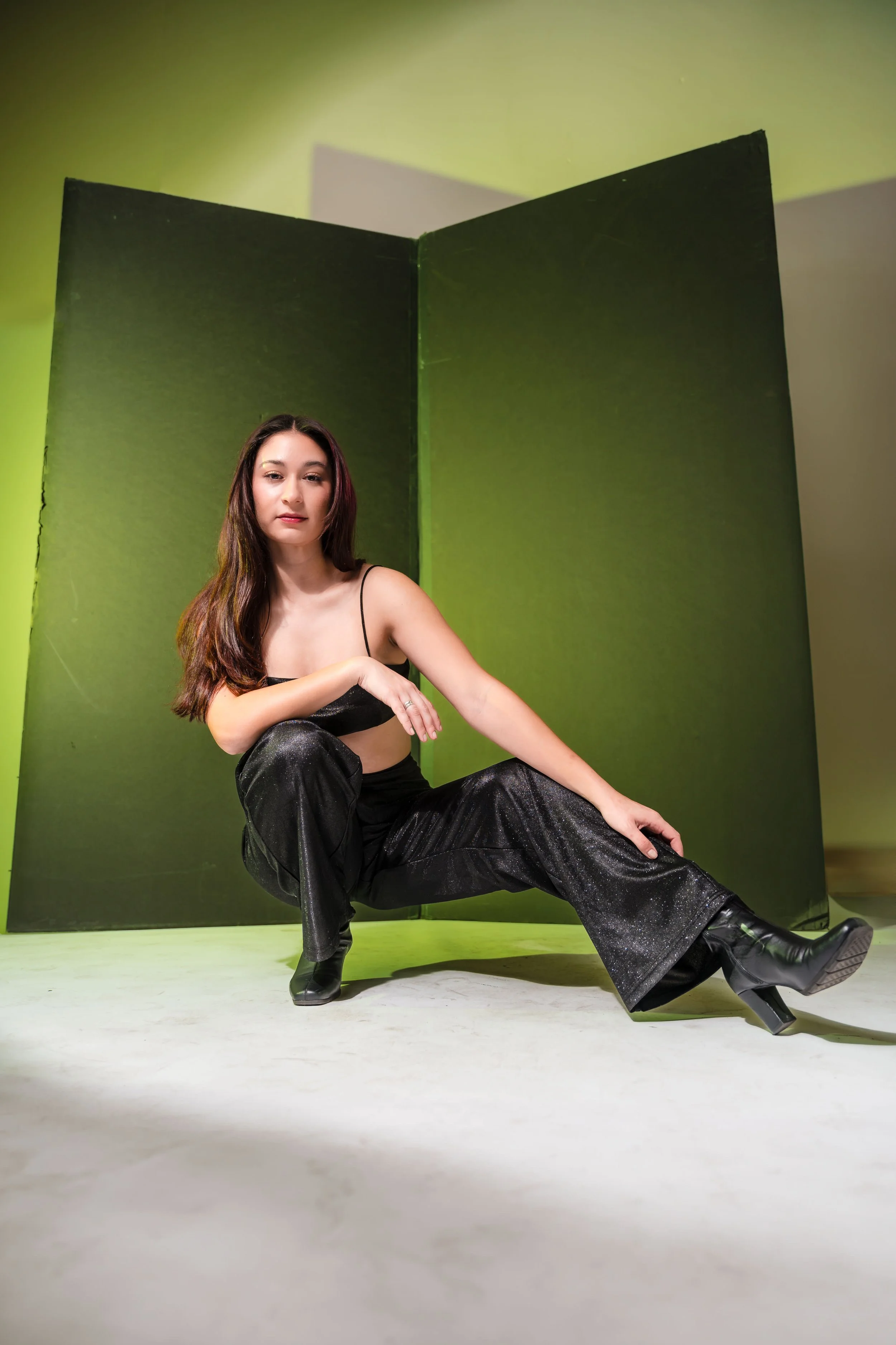 A woman with long dark hair in a black outfit sitting on the floor with one leg bent and the other stretched out. She is in front of a green background with a green folding screen behind her, and is wearing black boots.