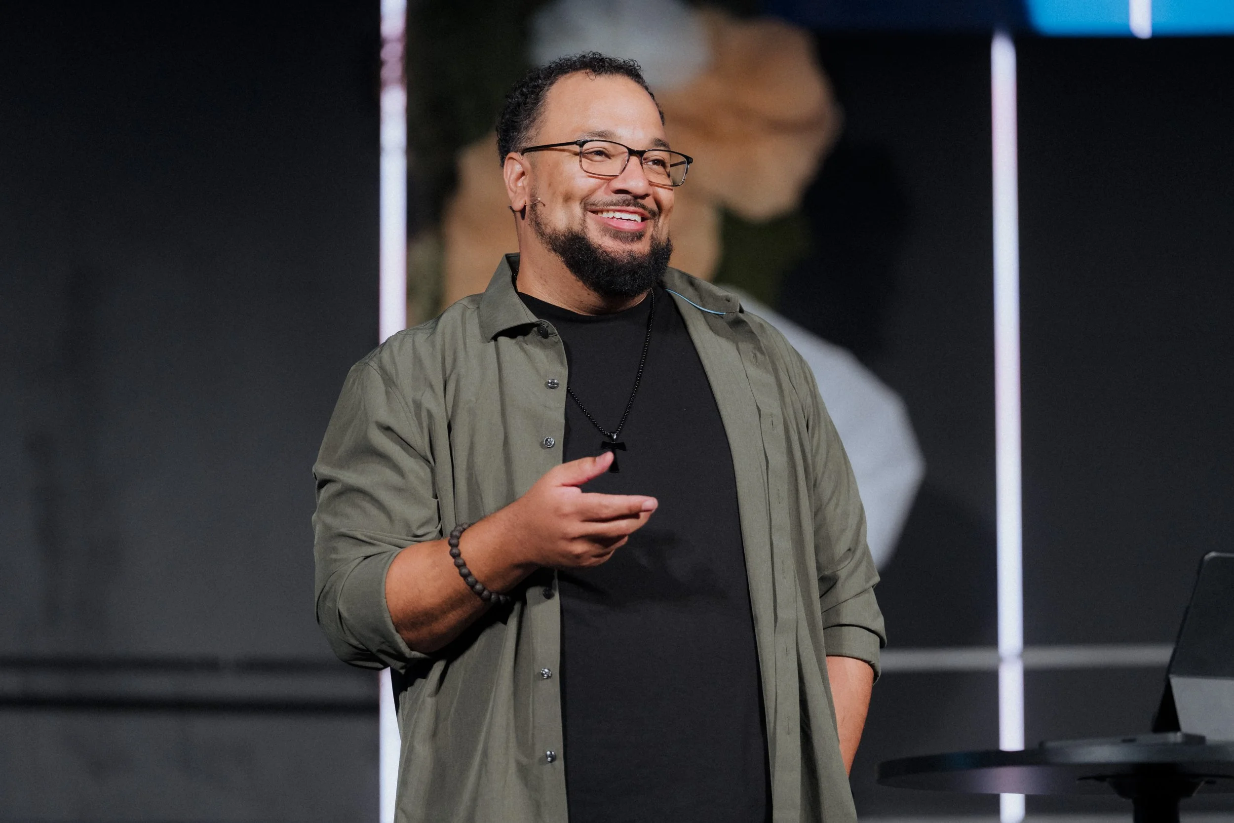 A man with glasses, a beard, and a goatee smiling while speaking at an event. He is wearing a black shirt, an open olive green button-up shirt, a beaded bracelet, and a necklace with a black pendant. He is standing near a table with a laptop on it, w