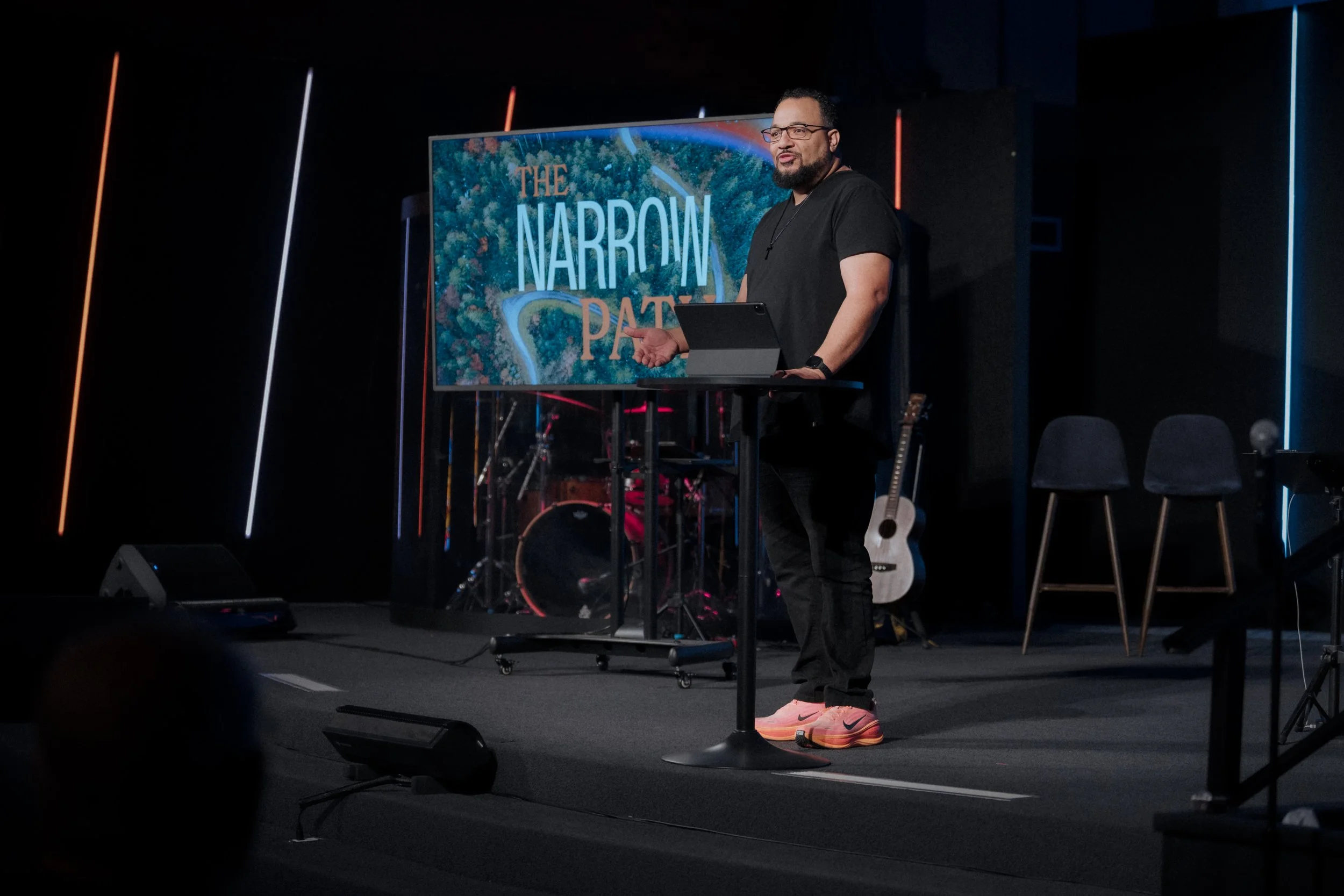 A man with glasses, beard, wearing a black shirt and pink Nike sneakers standing at a podium on a stage, gesturing with his hand, with a large screen behind him displaying the text 'The Narrow Path' and a forested landscape.