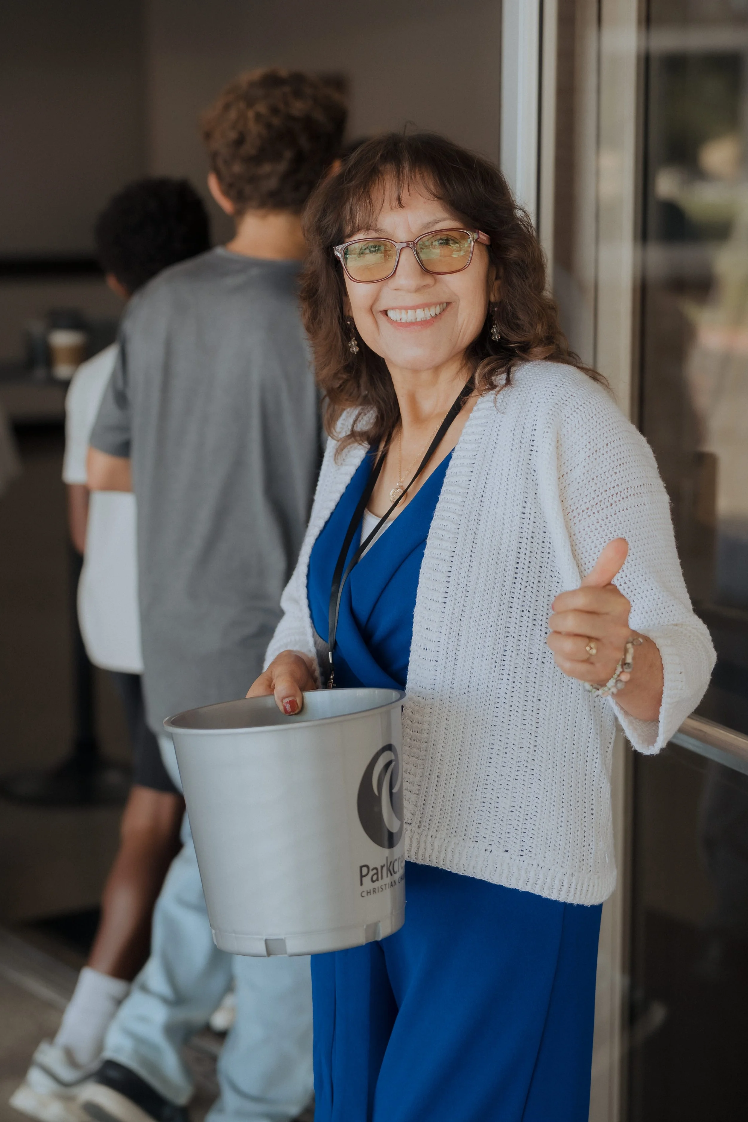 A woman wearing glasses, a white knit cardigan, and a blue dress is smiling and giving a thumbs-up while holding a metal collection bucket with a church logo on it. She is standing in front of a glass door with children lined up behind her.