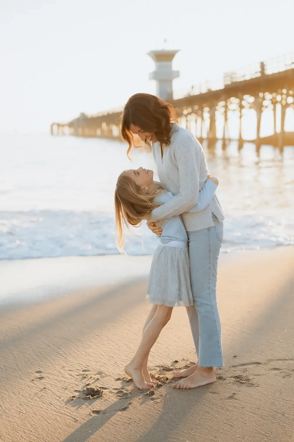 A mother and daughter sharing a joyful moment on a beach at sunset, with Seal Beach pier in the background.