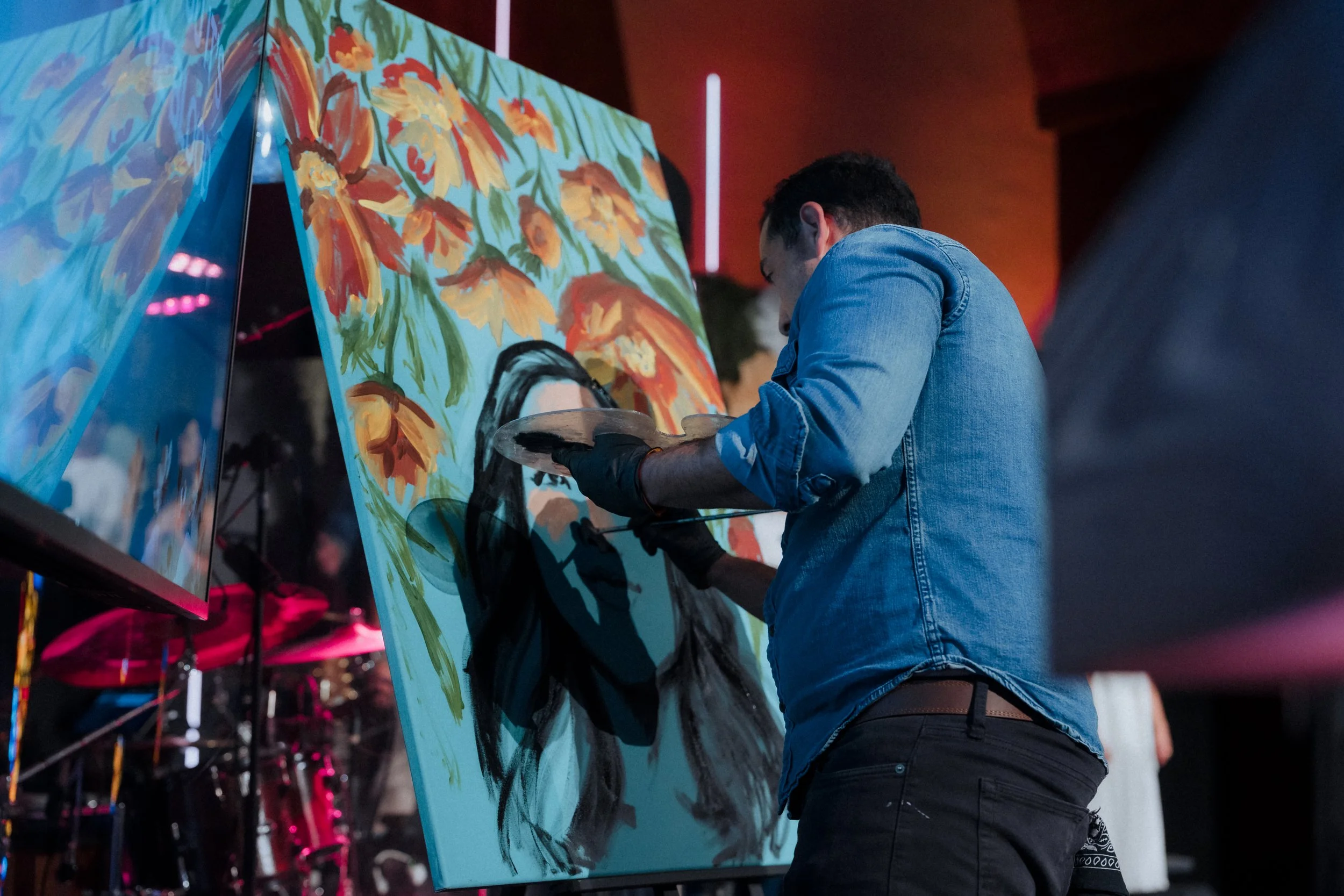Artist in blue denim jacket and black gloves painting a portrait of a woman on a large canvas with colorful floral background at an art event