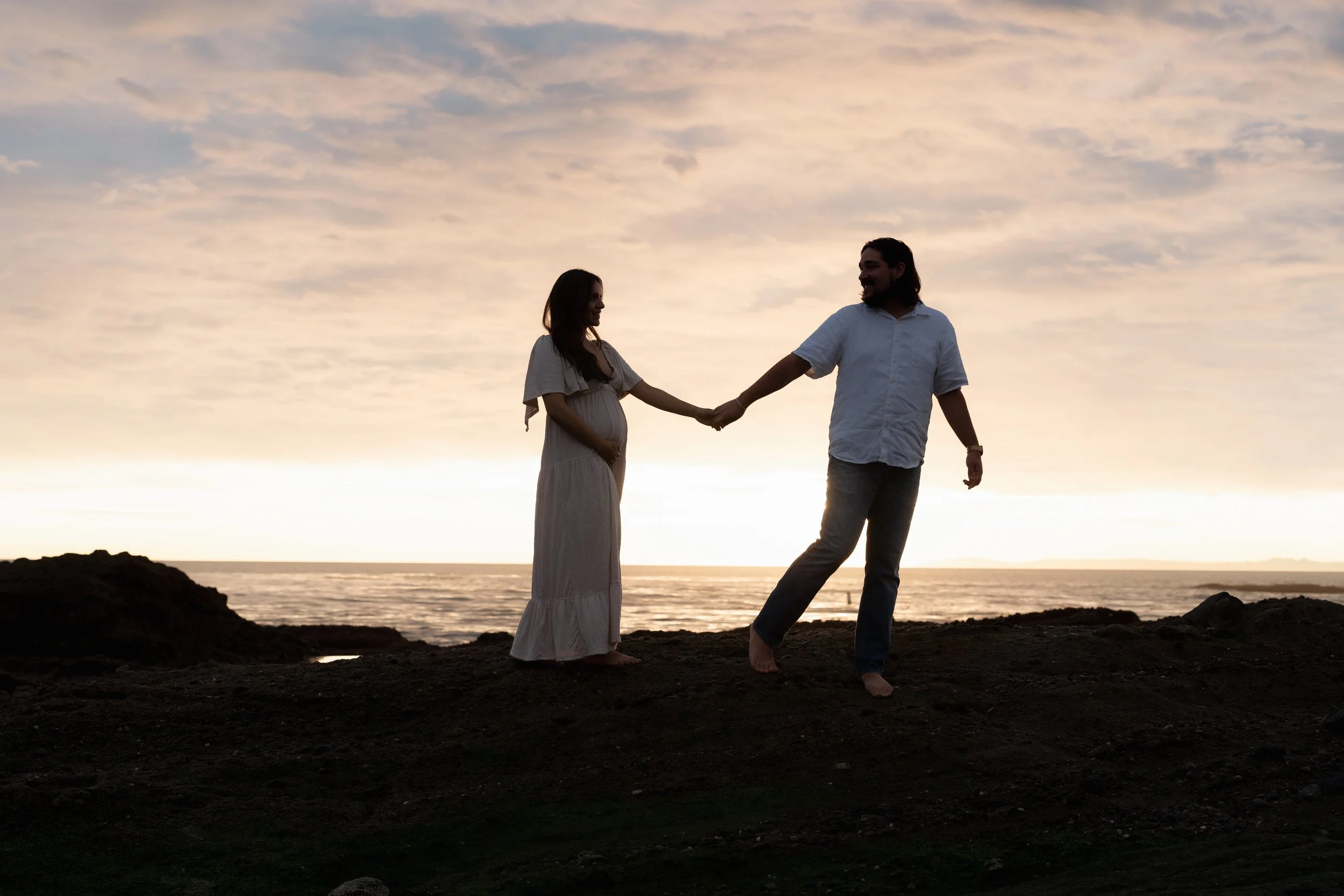A couple holding hands on a rocky beach at sunset, with the woman visibly pregnant and both smiling at each other.