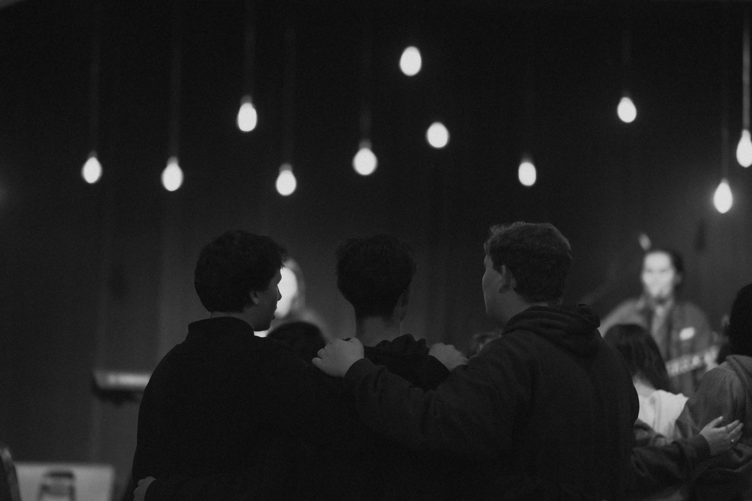 Three young men sitting with their arms around each other, watching a performance on stage in a dimly lit venue with hanging light bulbs overhead.