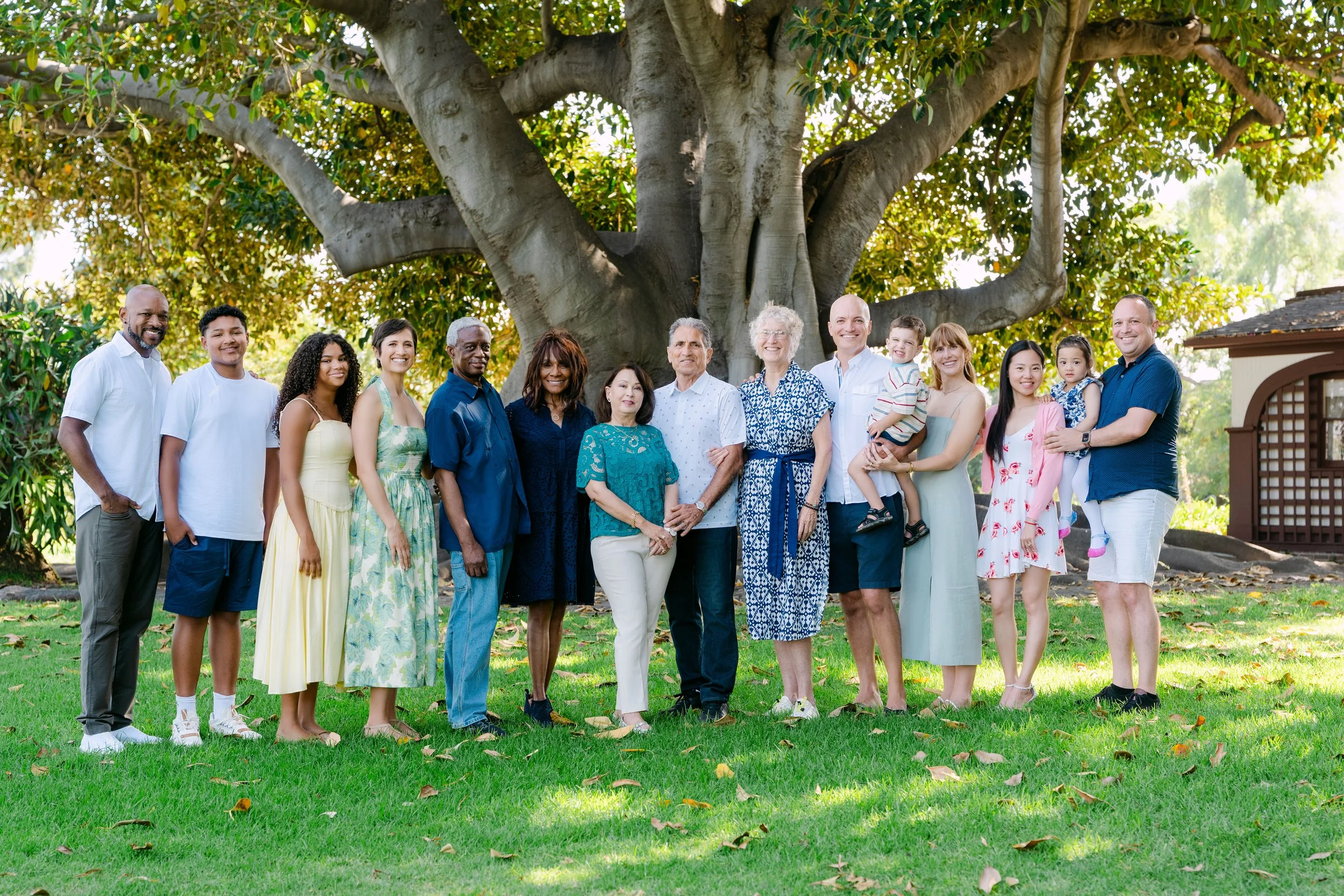 A diverse group of 15 people, including children and seniors, standing outdoors under a large tree, smiling at the camera.