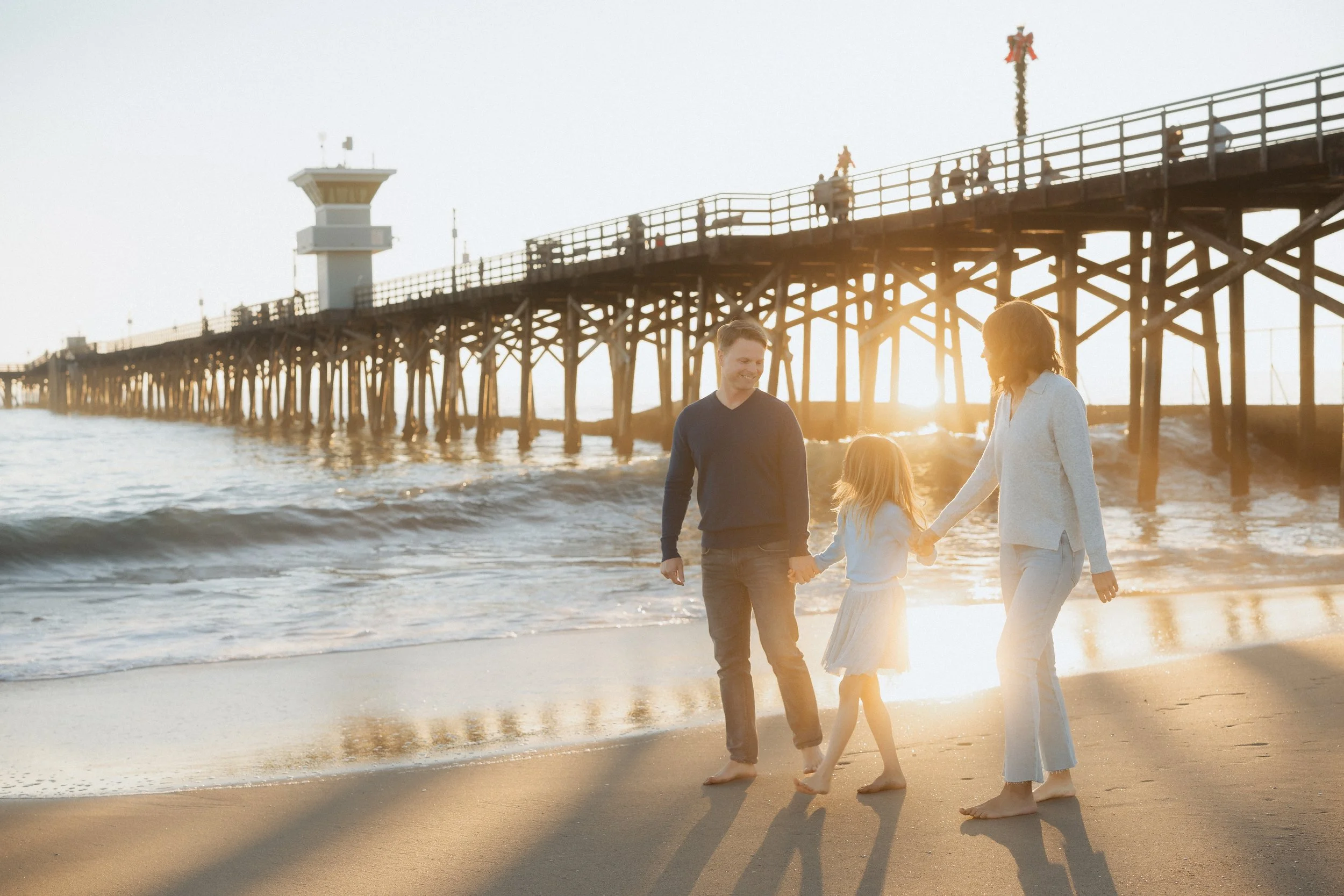 A family of three walking hand-in-hand along the beach at sunset, near a pier with a lighthouse structure.