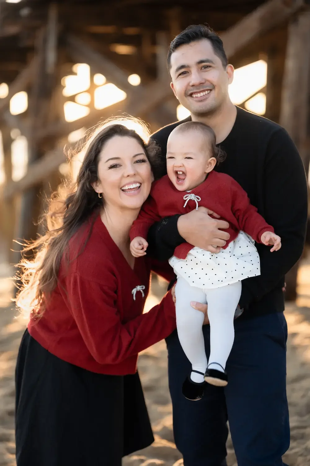 A happy family of three outdoors during sunset. The mother, father, and toddler girl are smiling and laughing. They are standing under a the wooden Seal Beach Pier.
