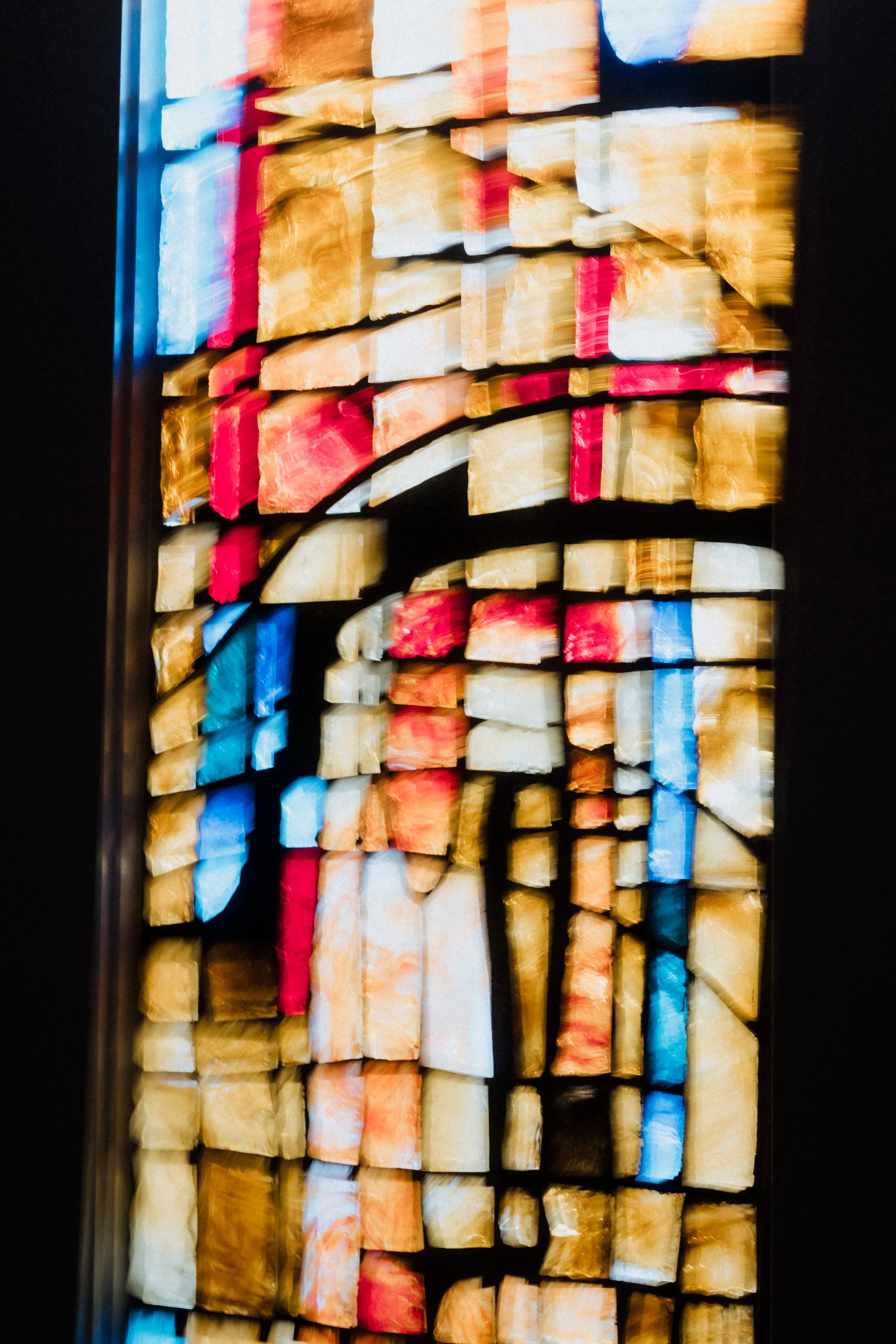 A colorful, abstract stained glass artwork made of rectangular and irregular glass pieces in shades of gold, red, blue, white, and black.