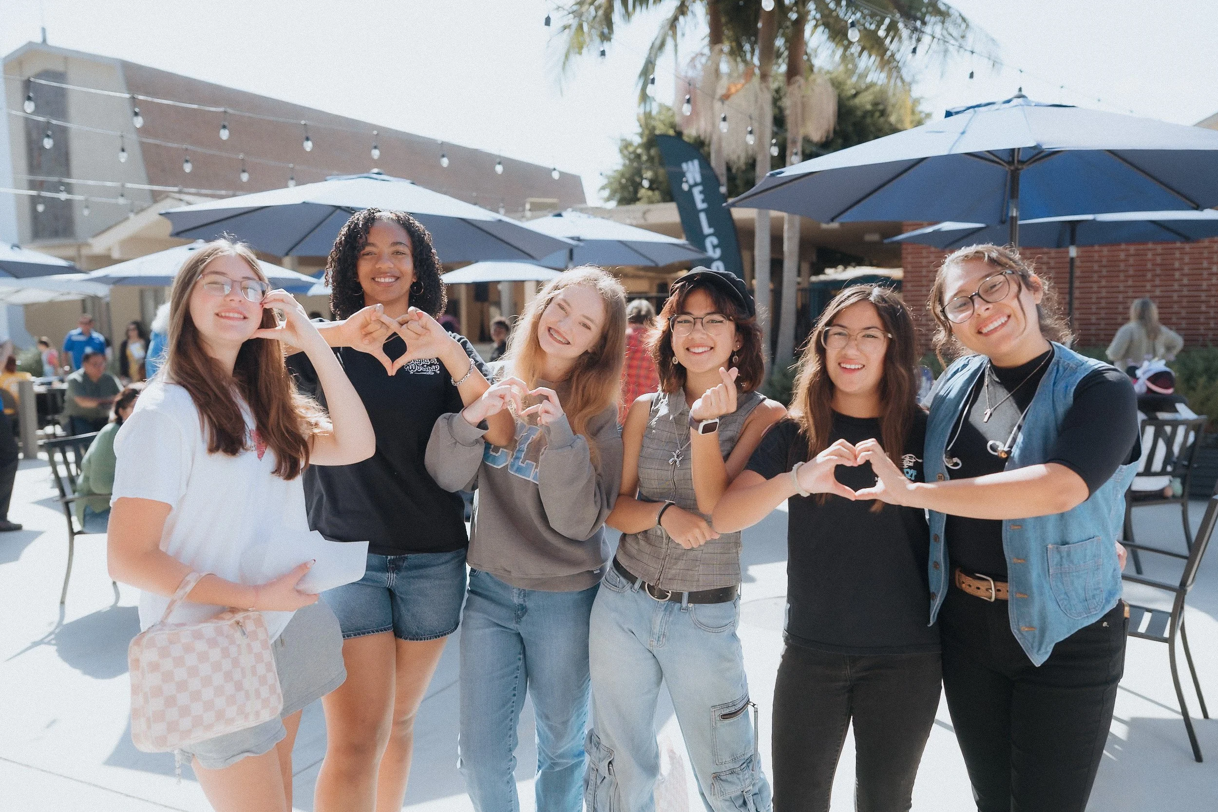 Six young women standing outdoors at a social gathering, making heart shapes with their hands, with tables, umbrellas, and people in the background.