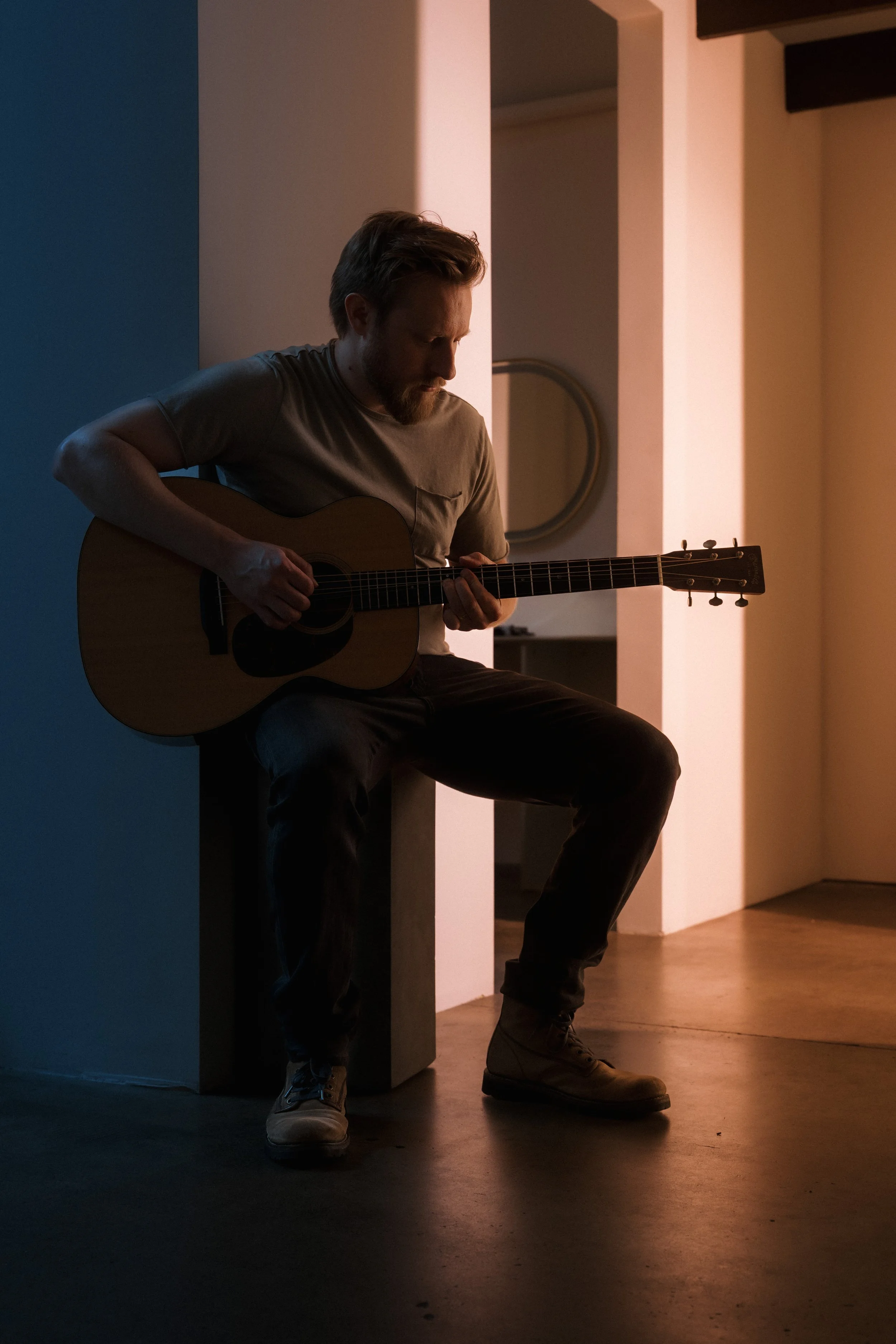 A man with a beard playing an acoustic guitar while sitting on a ledge in a dimly lit room with modern decor.