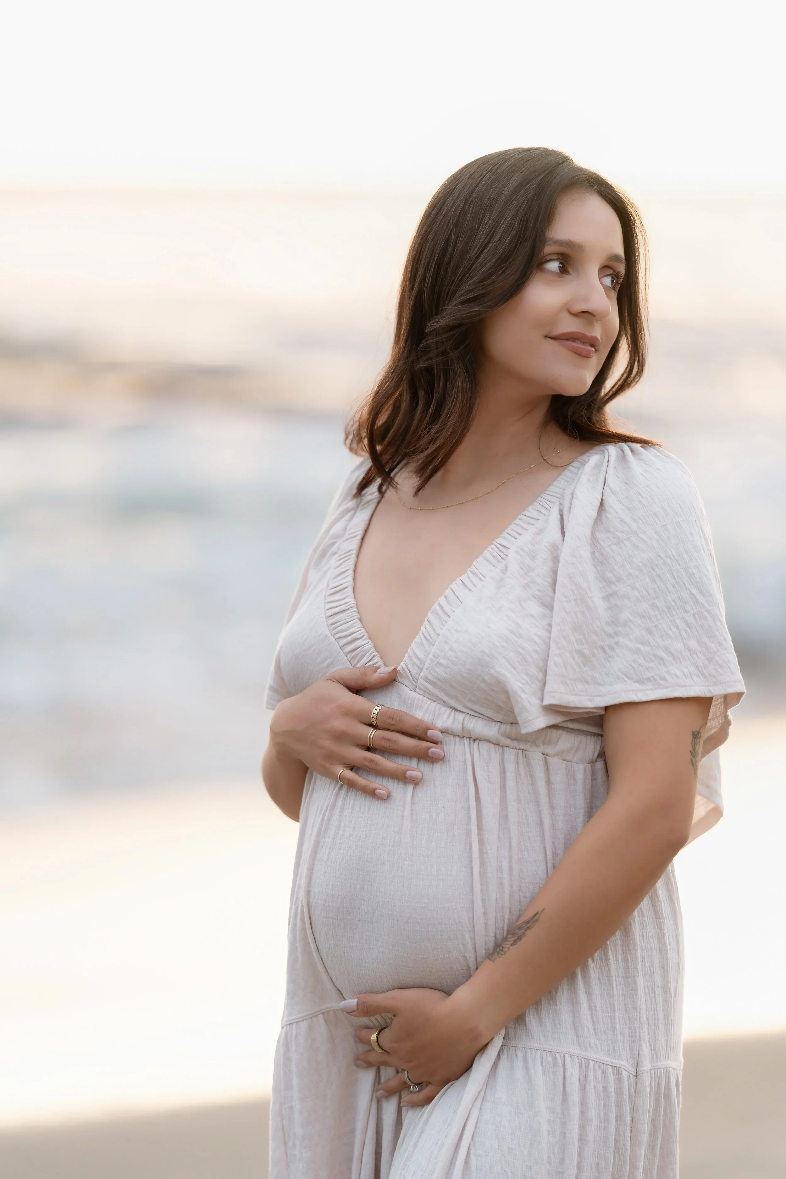 A pregnant woman standing on the beach with ocean waves in the background, wearing a light-colored dress and rings, looking to the side.