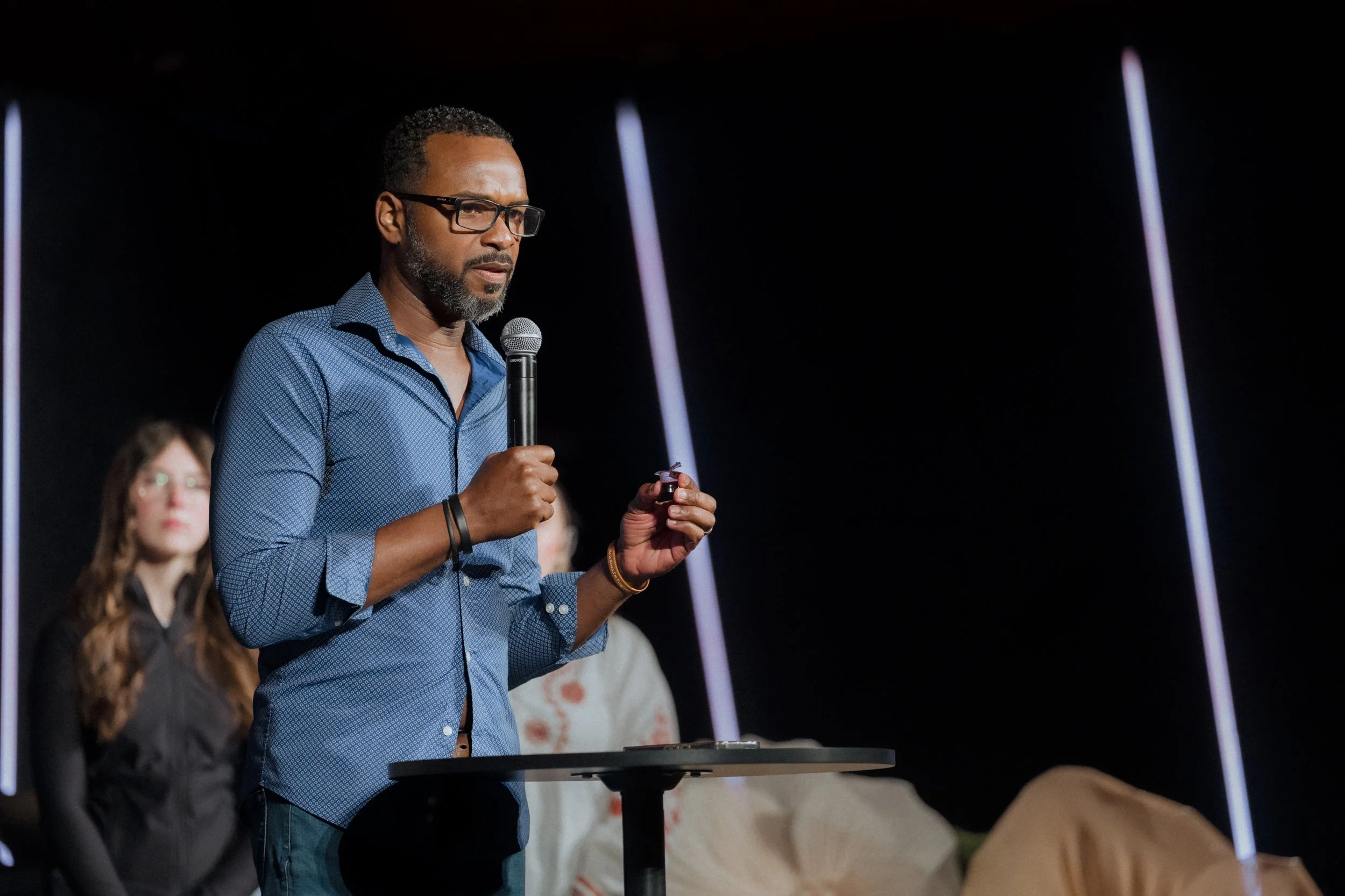 A man with glasses and a beard, wearing a blue shirt, is holding a microphone and speaking at a presentation or conference. Two women are standing in the background, and the setting appears to be a dark auditorium or stage.
