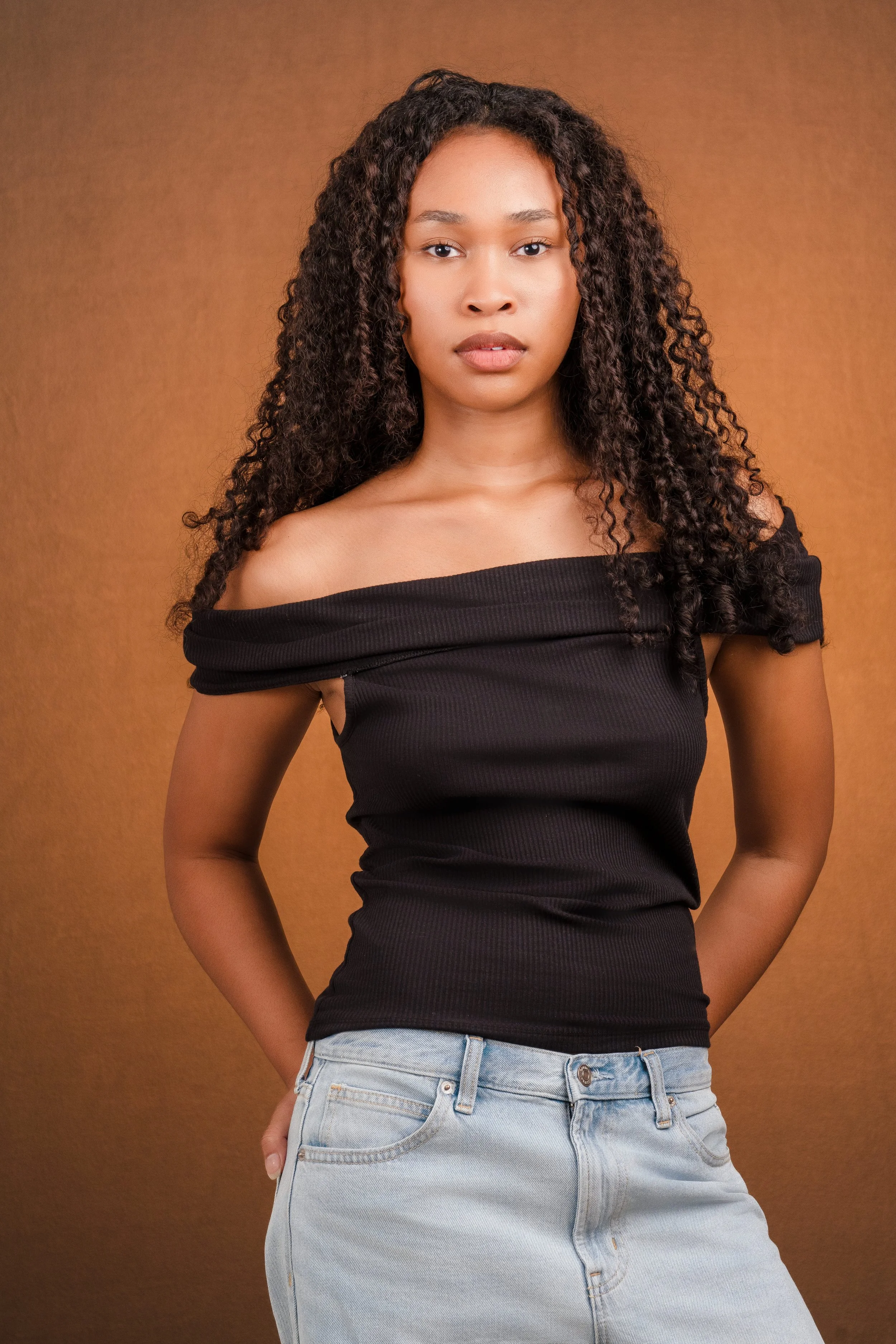 A young woman with curly hair wearing a black off-the-shoulder top and light blue jeans standing against a brown background.